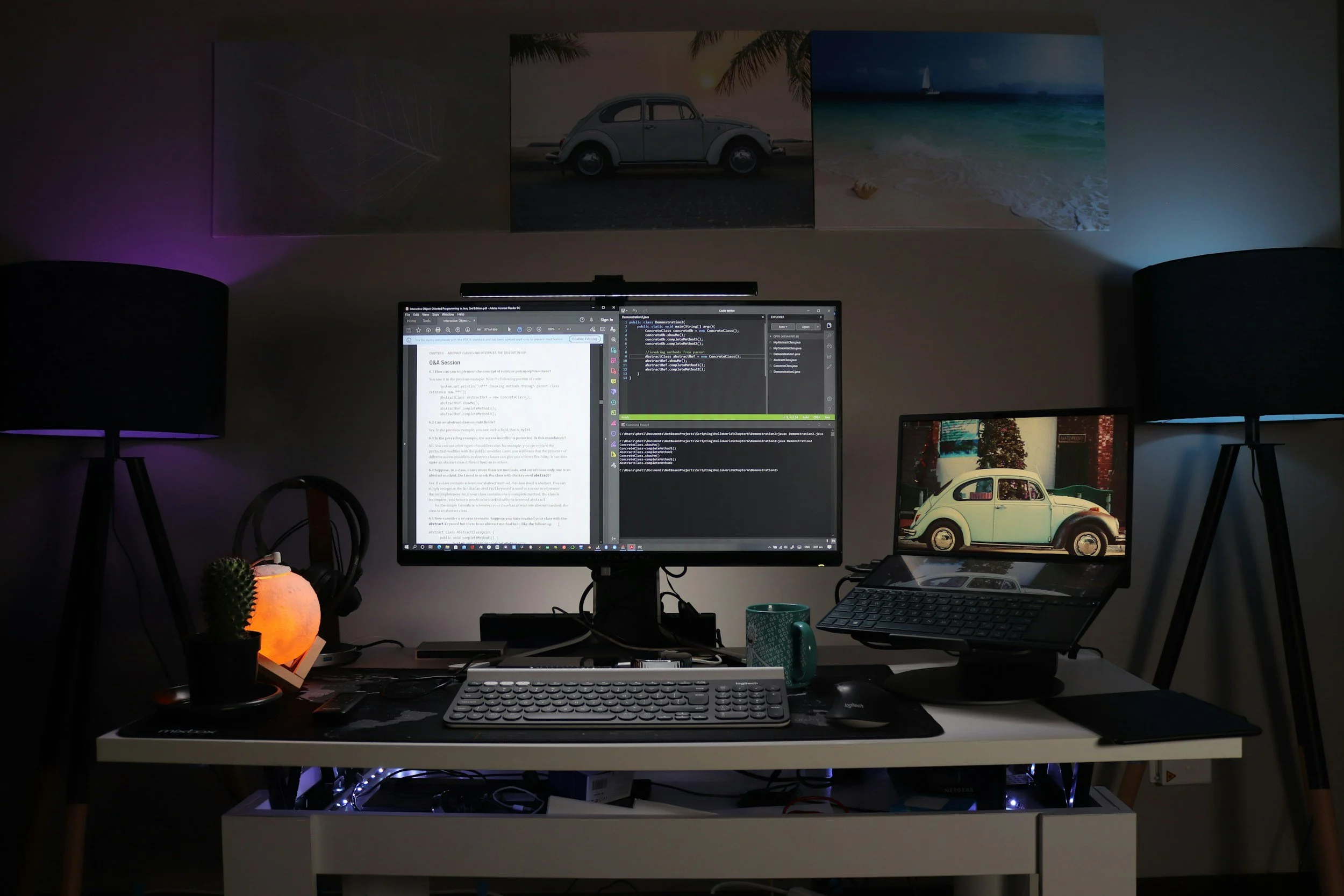 Estação de trabalho com dois monitores, teclado, mouse e iluminação suave, decoração com quadros na parede, incluindo uma imagem de um carro antigo e uma praia, ao lado de duas luminárias de mesa acesas.