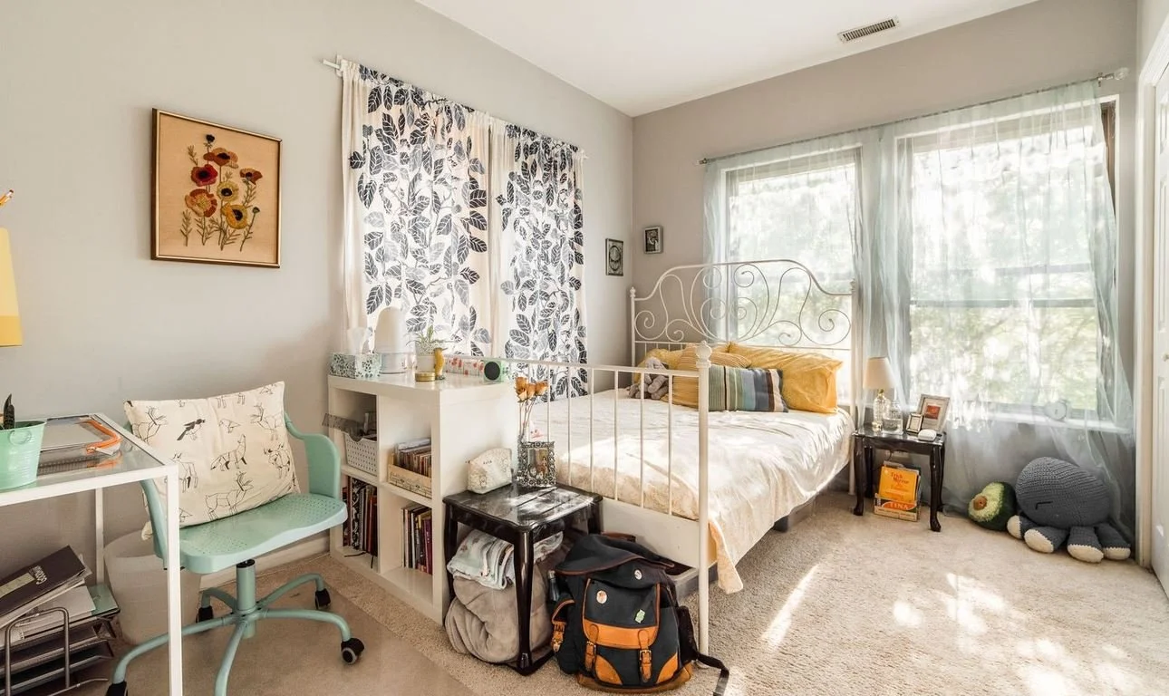 Bedroom with large window, white metal bed frame, yellow pillows, black nightstand with a table lamp, plush toy, and framed photos, dresser with books and small decorations, desk with green chair, floral curtains, and wall art of flowers.