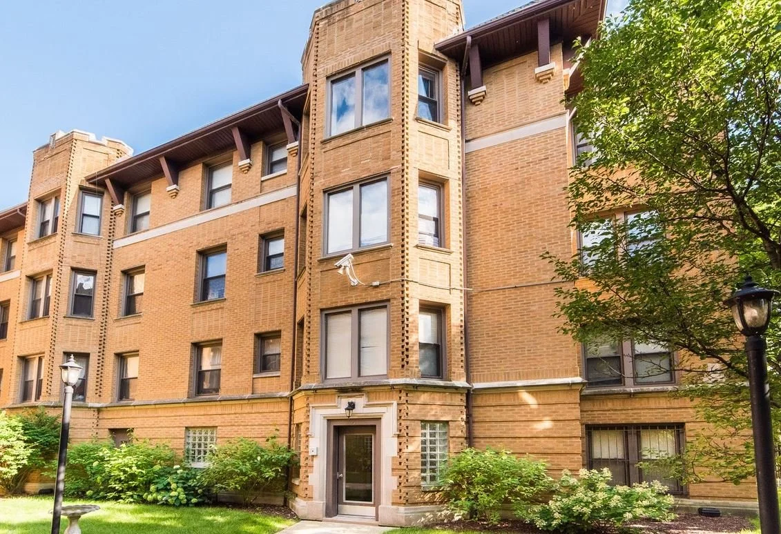 A multi-story brick apartment building with several windows, a main entrance with a glass door, surrounded by greenery including bushes, trees, and lawn, with street lamps in the foreground.