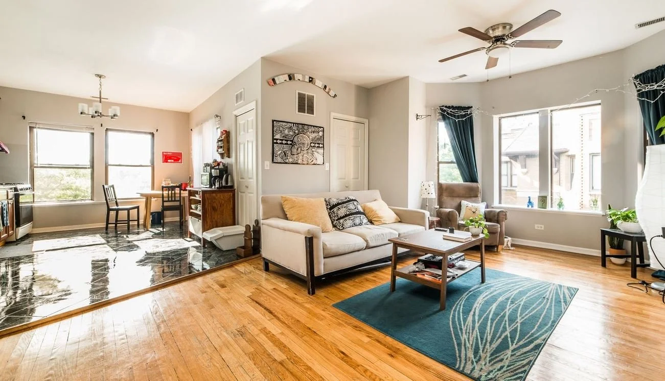 Open-concept living room with hardwood floors, a cream sofa with pillows, a blue area rug, a coffee table, large windows with curtains, and a ceiling fan.