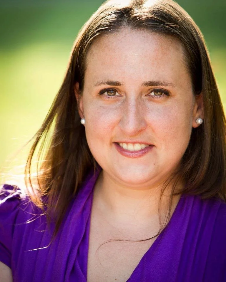 A young woman with brown hair, wearing pearl earrings and a purple top, smiling outdoors with green blurred background.