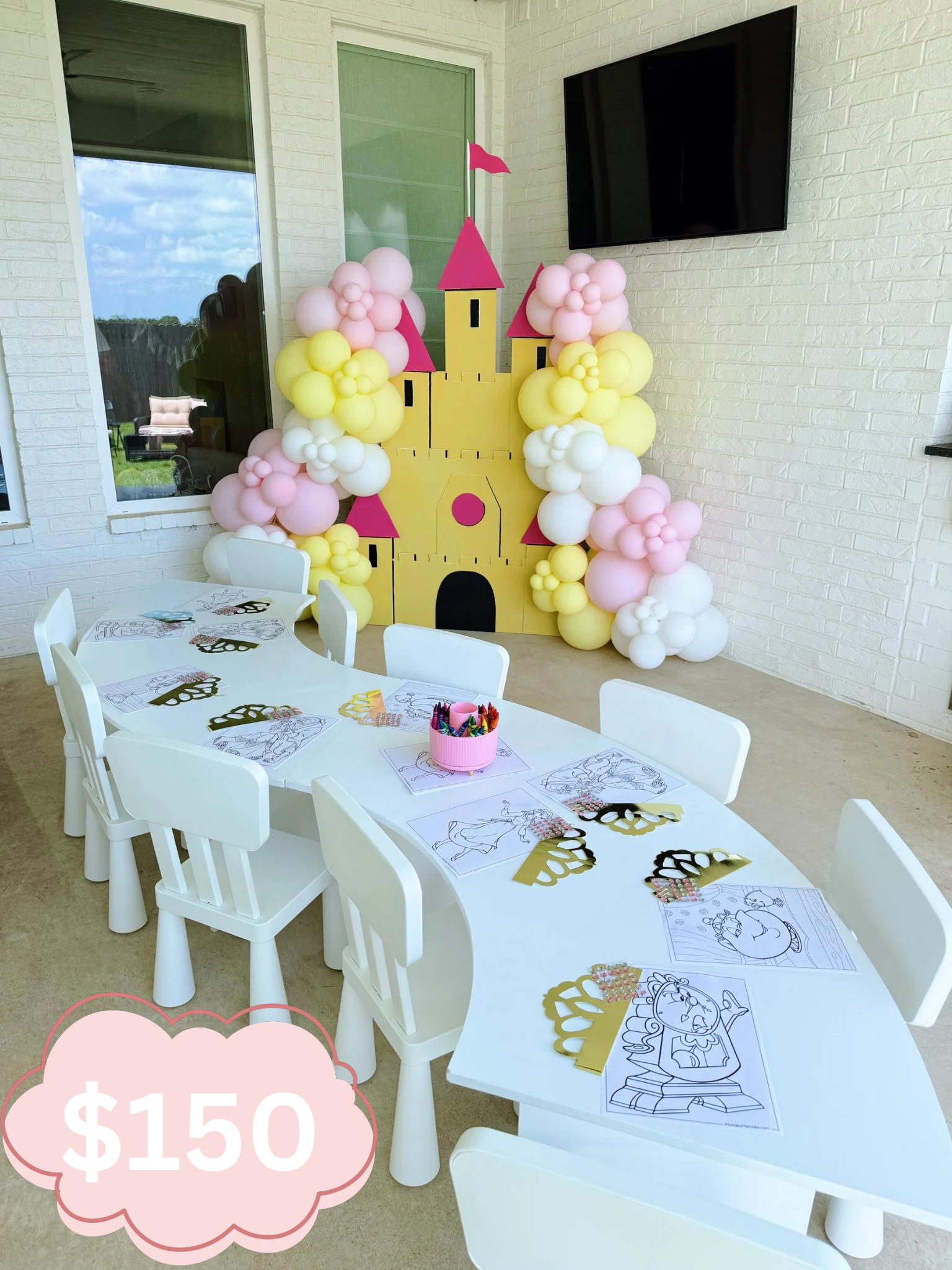 Party setup featuring a large castle-shaped balloon decoration with pink, yellow, and white balloons, pink castle towers, and yellow castle walls, with tables ready for children to color princess-themed coloring pages, surrounded by party accessories and decorations, in a bright indoor setting with windows and a wall-mounted TV.