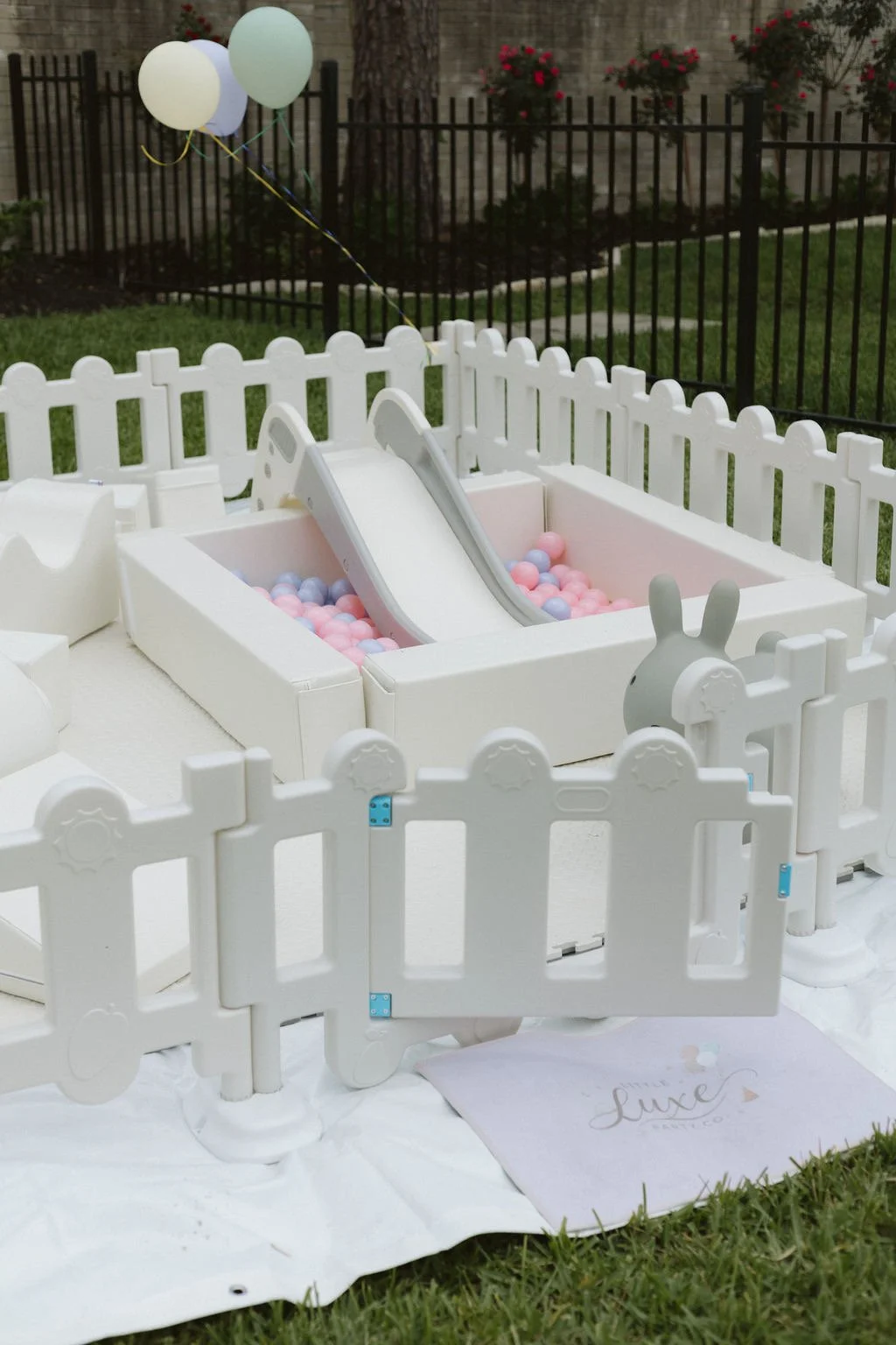 Small white plastic playpen with a slide leading into a ball pit filled with pink, blue, and purple balls, outdoors on grass with a white cloth underneath.