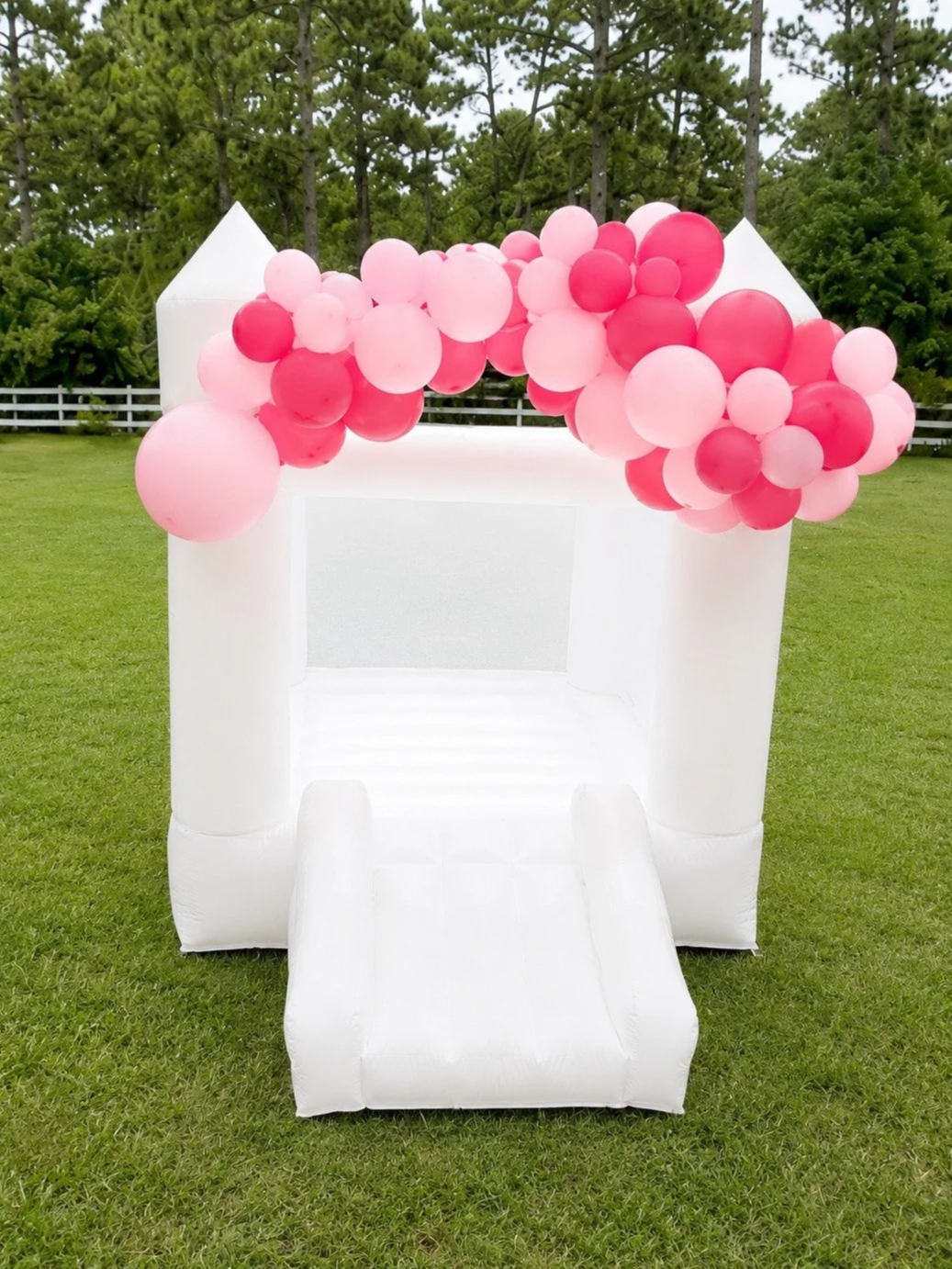 White inflatable photo booth decorated with a pink and red balloon arch, set up outdoors on green grass with trees in the background.