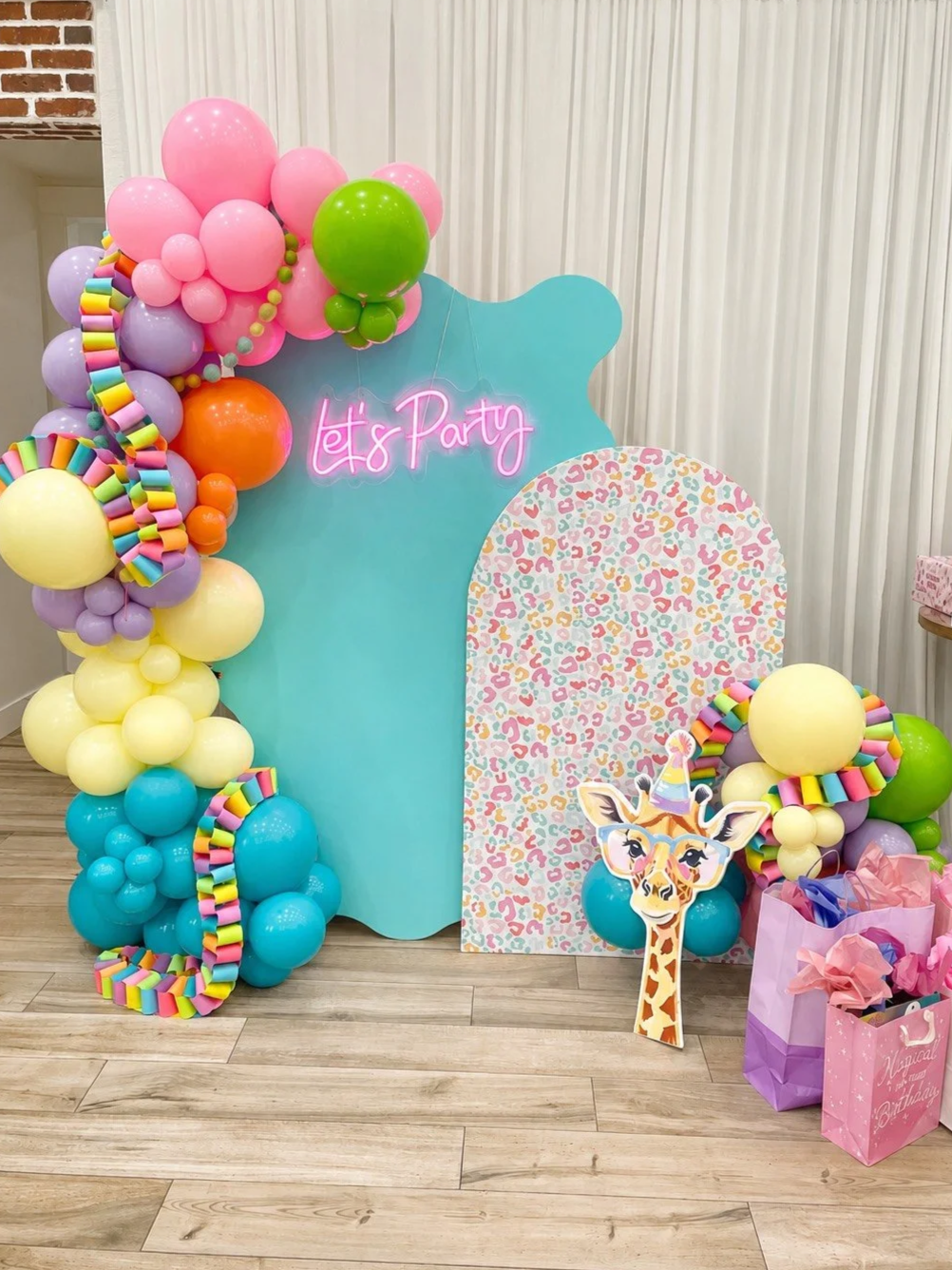 Colorful birthday party backdrop with balloons, a pink neon sign that reads 'Let's Party,' and a giraffe cutout wearing a party hat. Gift bags are on the floor next to the backdrop.
