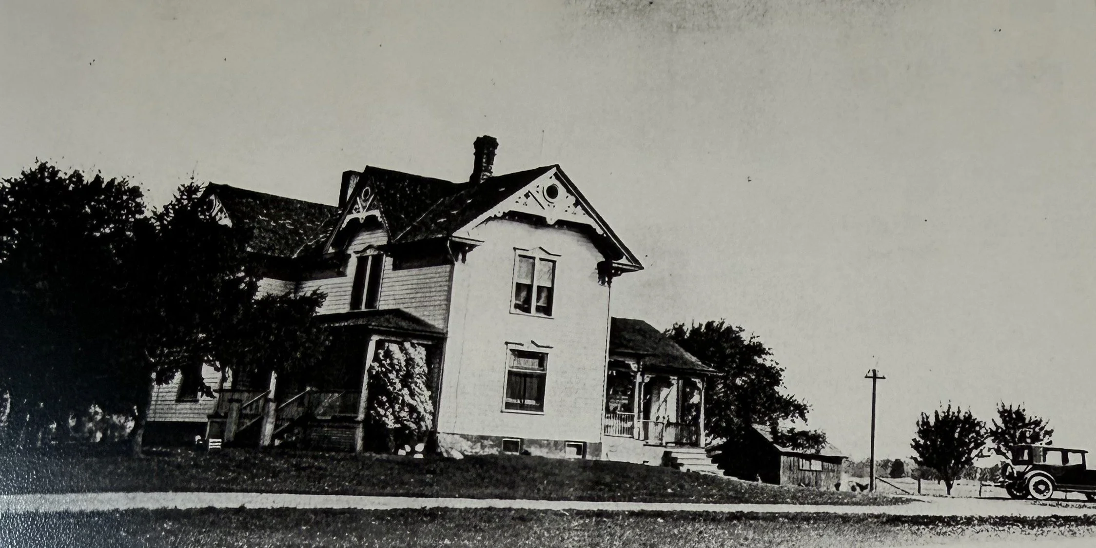 A black and white photograph of a two-story house with a porch, trees, and a vintage car parked on the street.