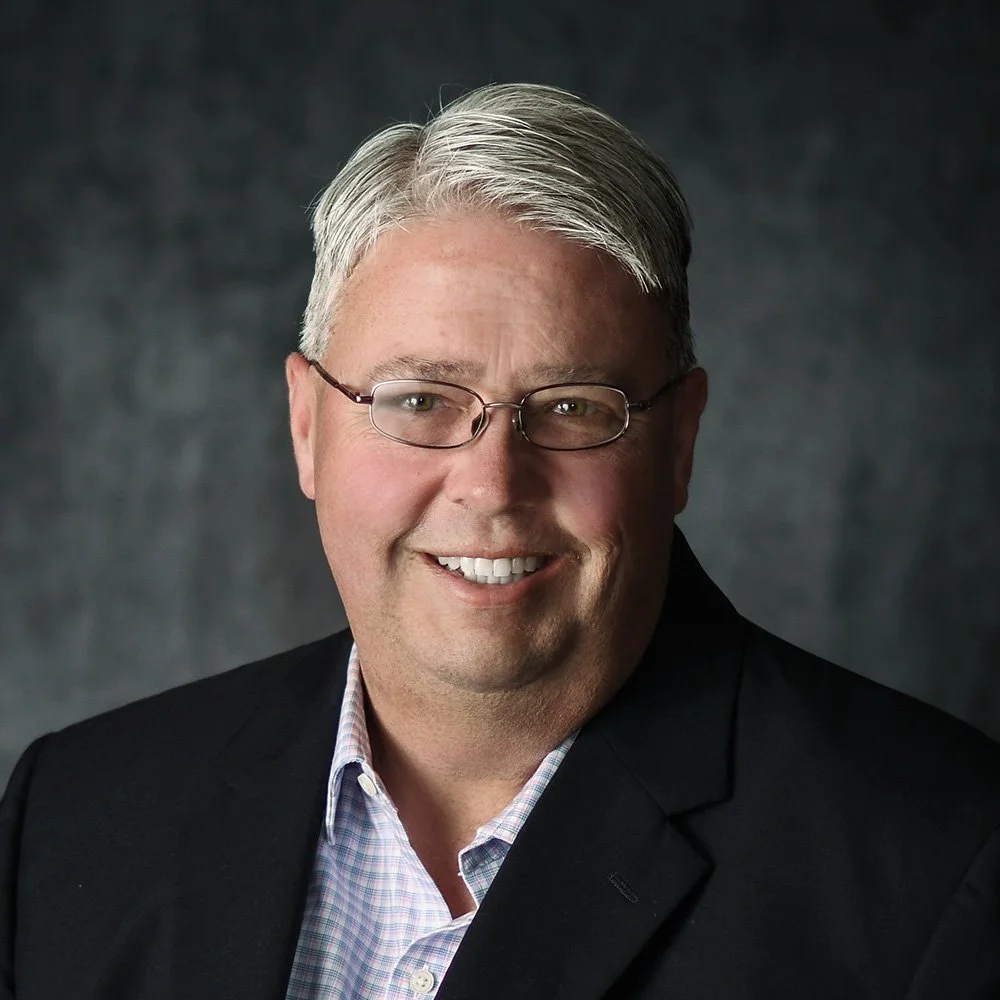 Smiling man with gray hair, glasses, wearing a black blazer and light checkered shirt, against a dark gray background.