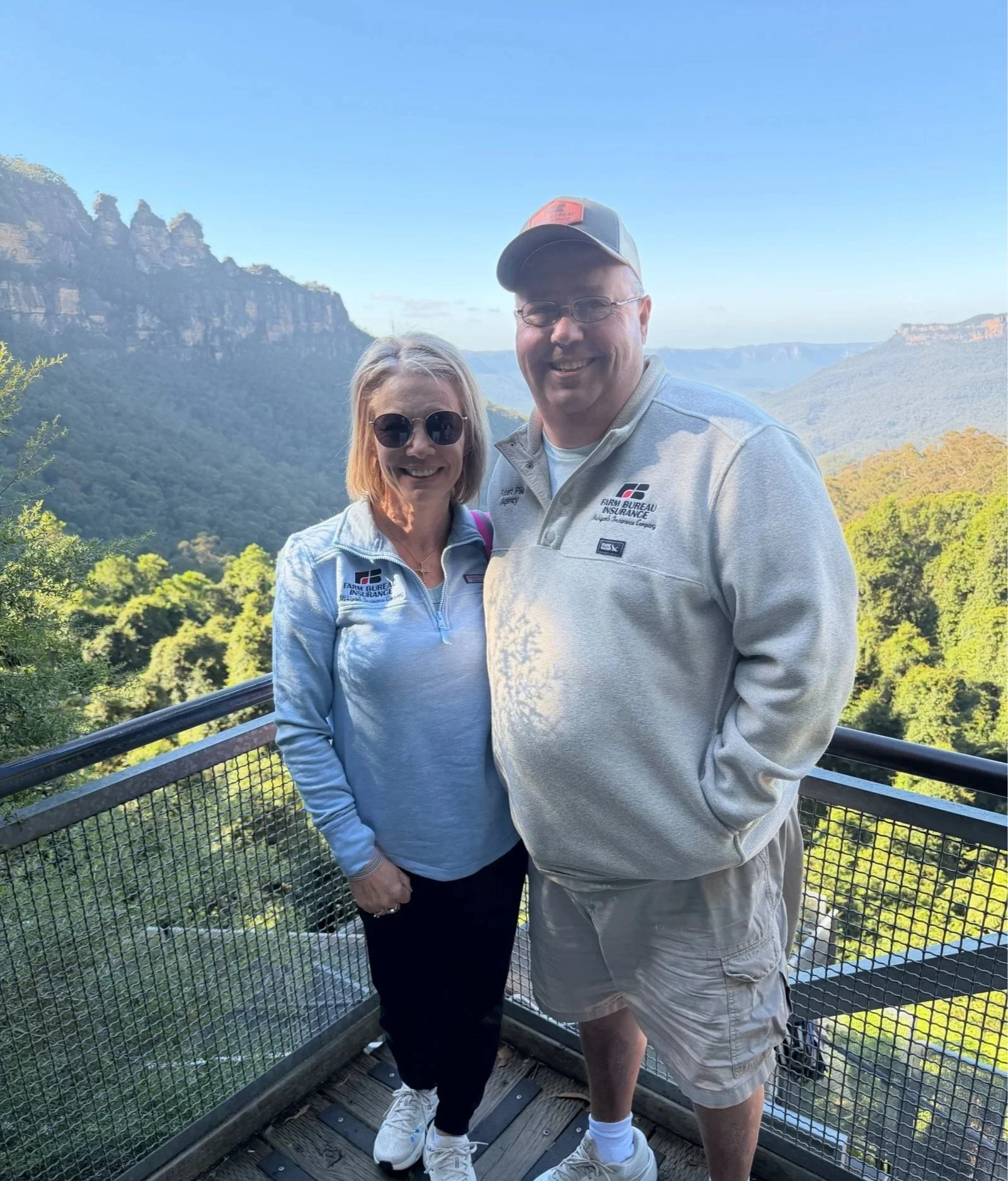 A smiling couple standing on a viewing platform with a scenic mountain landscape in the background. The woman is wearing sunglasses, a light gray jacket with a logo, and dark pants. The man is wearing glasses, a gray cap, a light gray quarter-zip pullover with a logo, and khaki shorts.