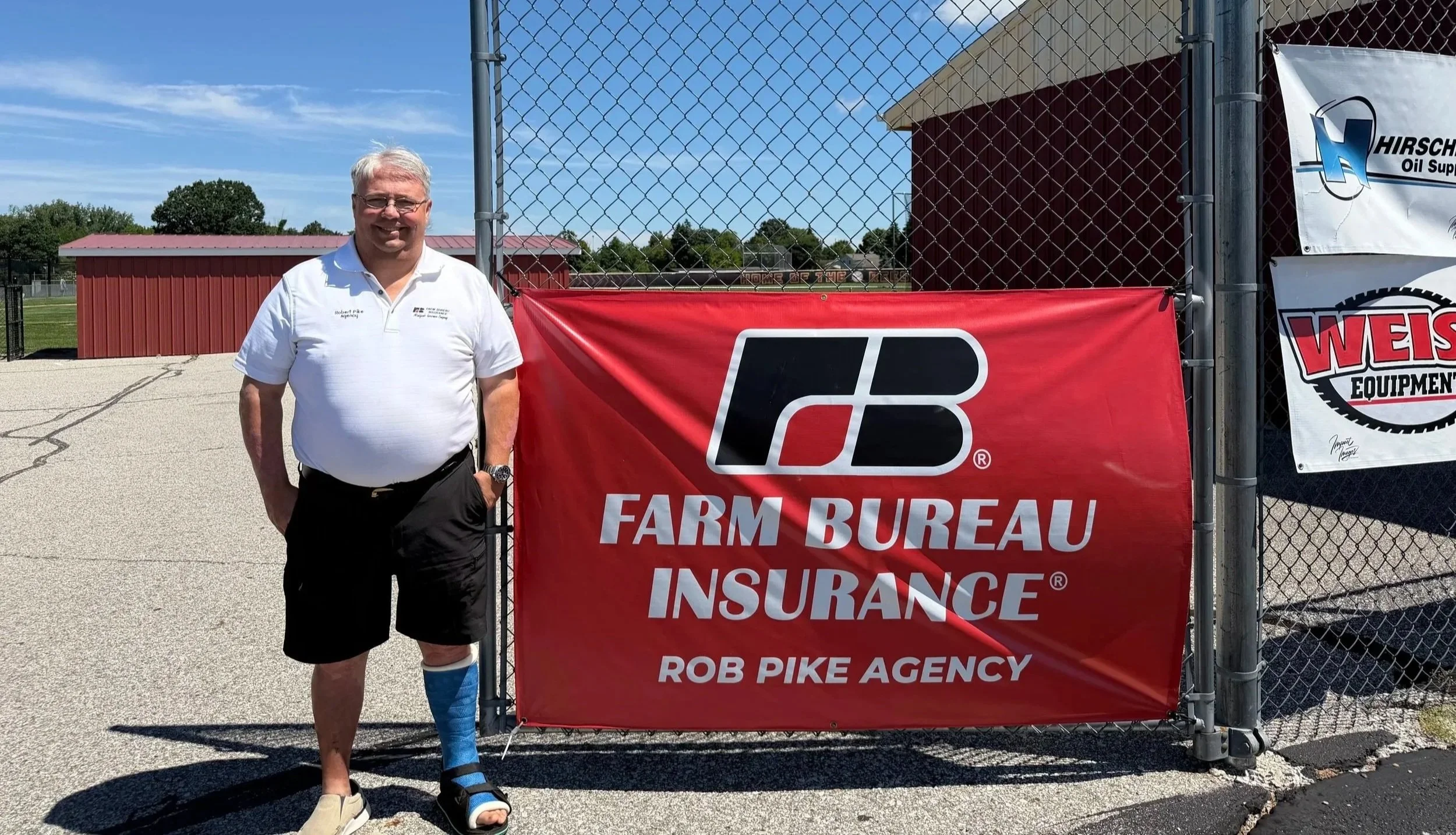 Man standing next to a red banner for Farm Bureau Insurance at a baseball field. The man has a bandage on his left leg and is smiling, wearing a white polo shirt and black shorts.
