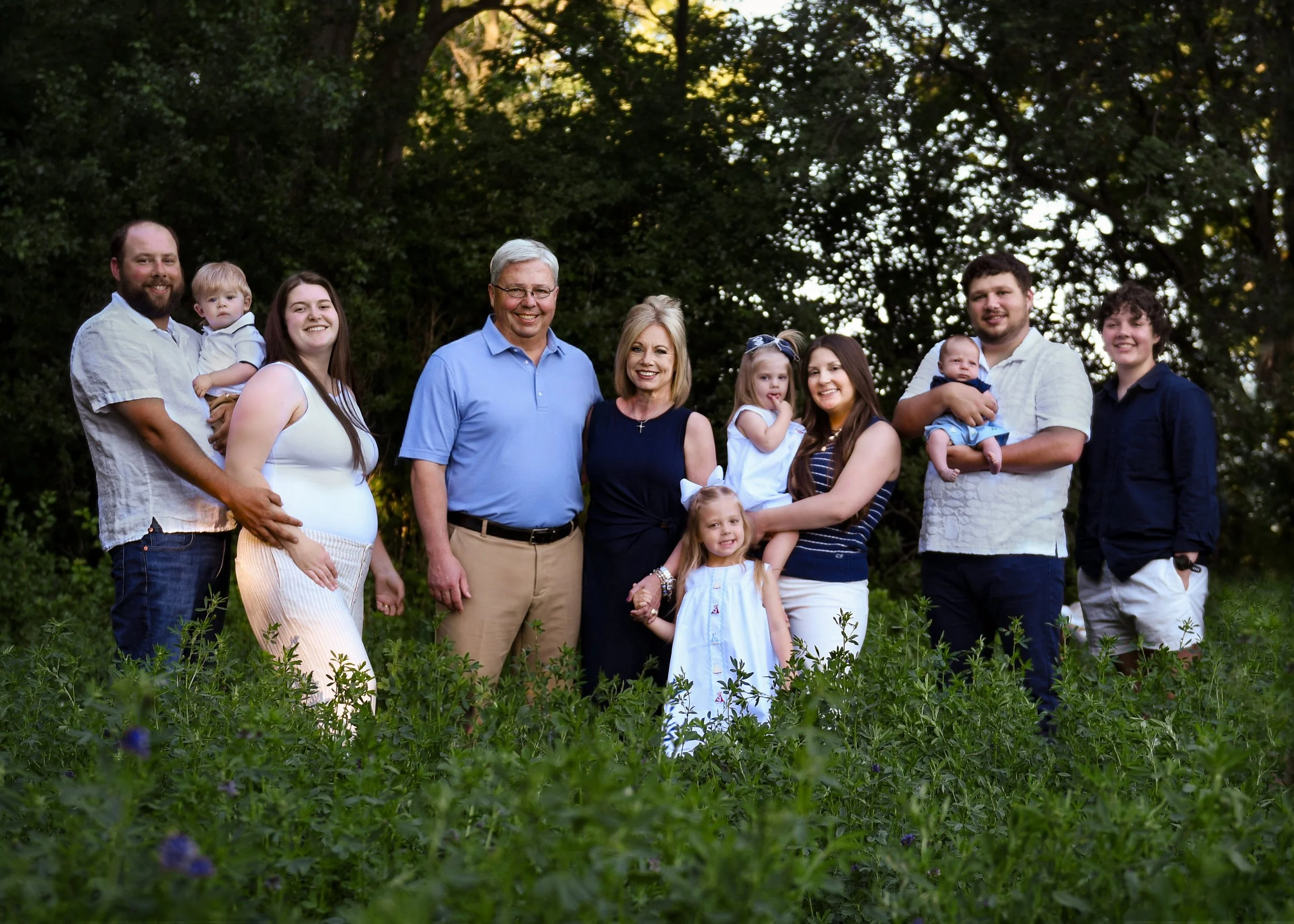 A multi-generational family standing outdoors in a lush, green setting with trees in the background, smiling at the camera.