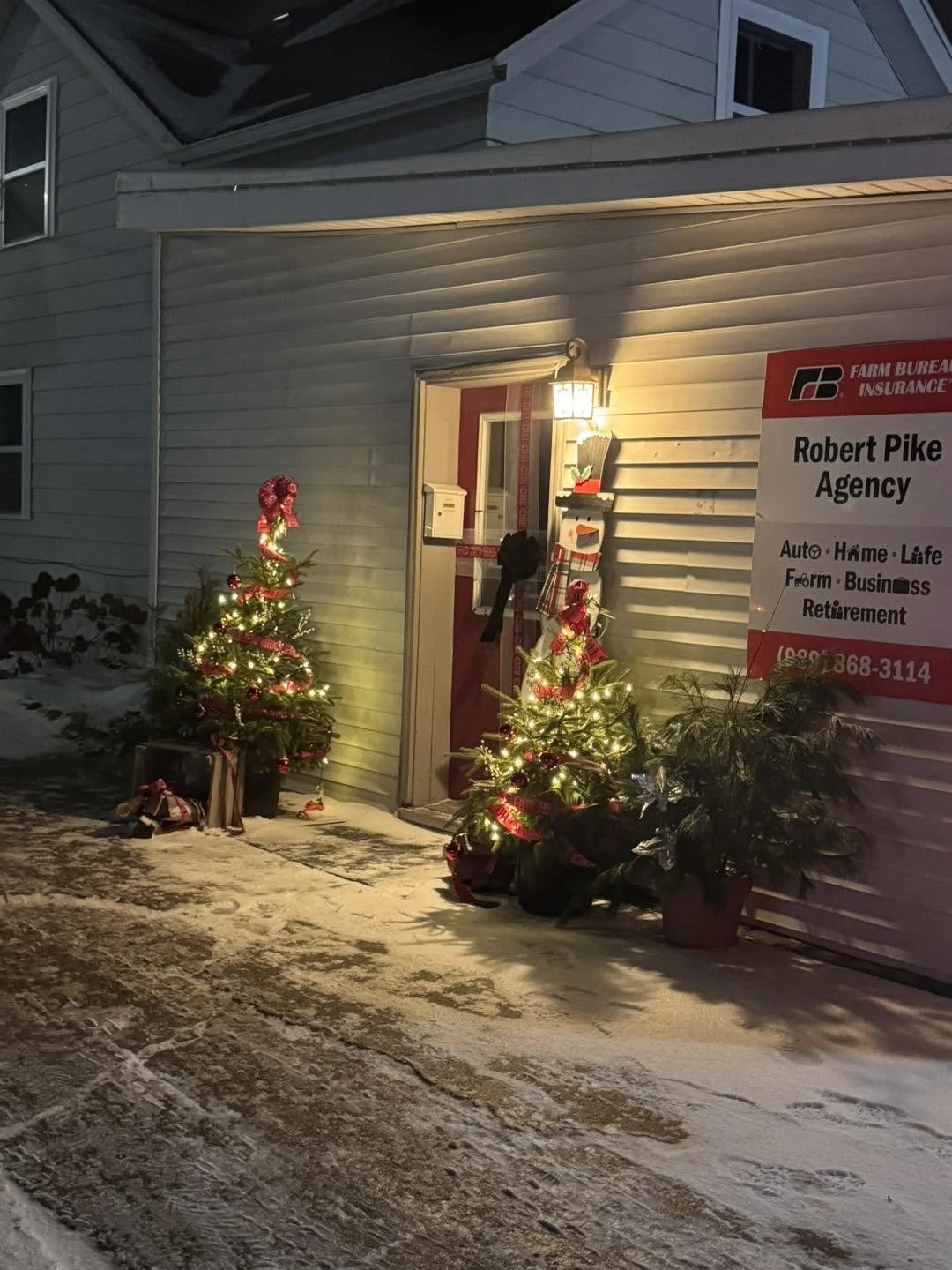 Outdoor scene with two decorated Christmas trees with lights and ribbons, placed on snow-covered ground in front of a white house. The house has a porch light on, and a red door with window and mailbox. Potted plants and a sign for Robert Pike Agency are visible on the porch.