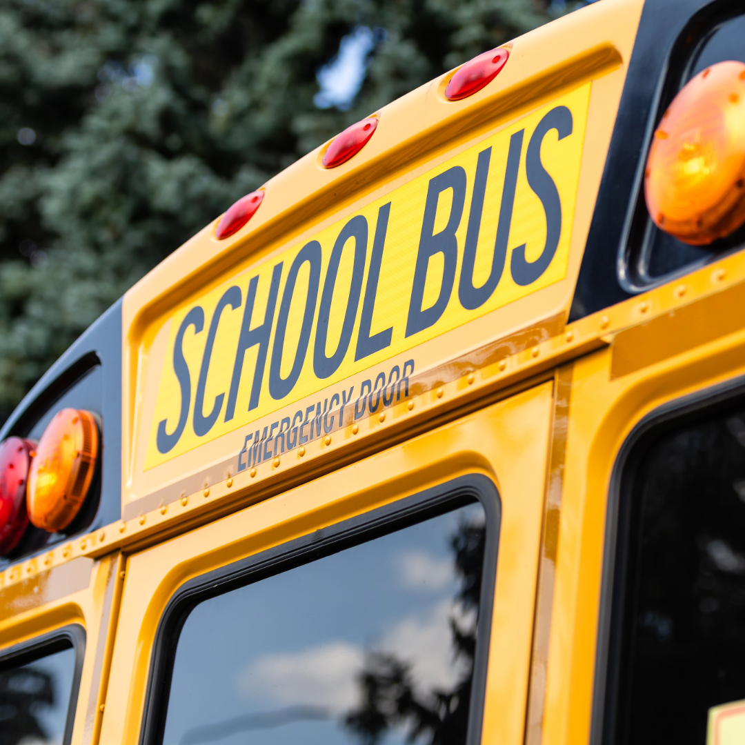 Close-up of the rear of a yellow school bus with a sign that reads 'SCHOOL BUS' and 'EMERGENCY DOOR' below it, including red and amber warning lights.