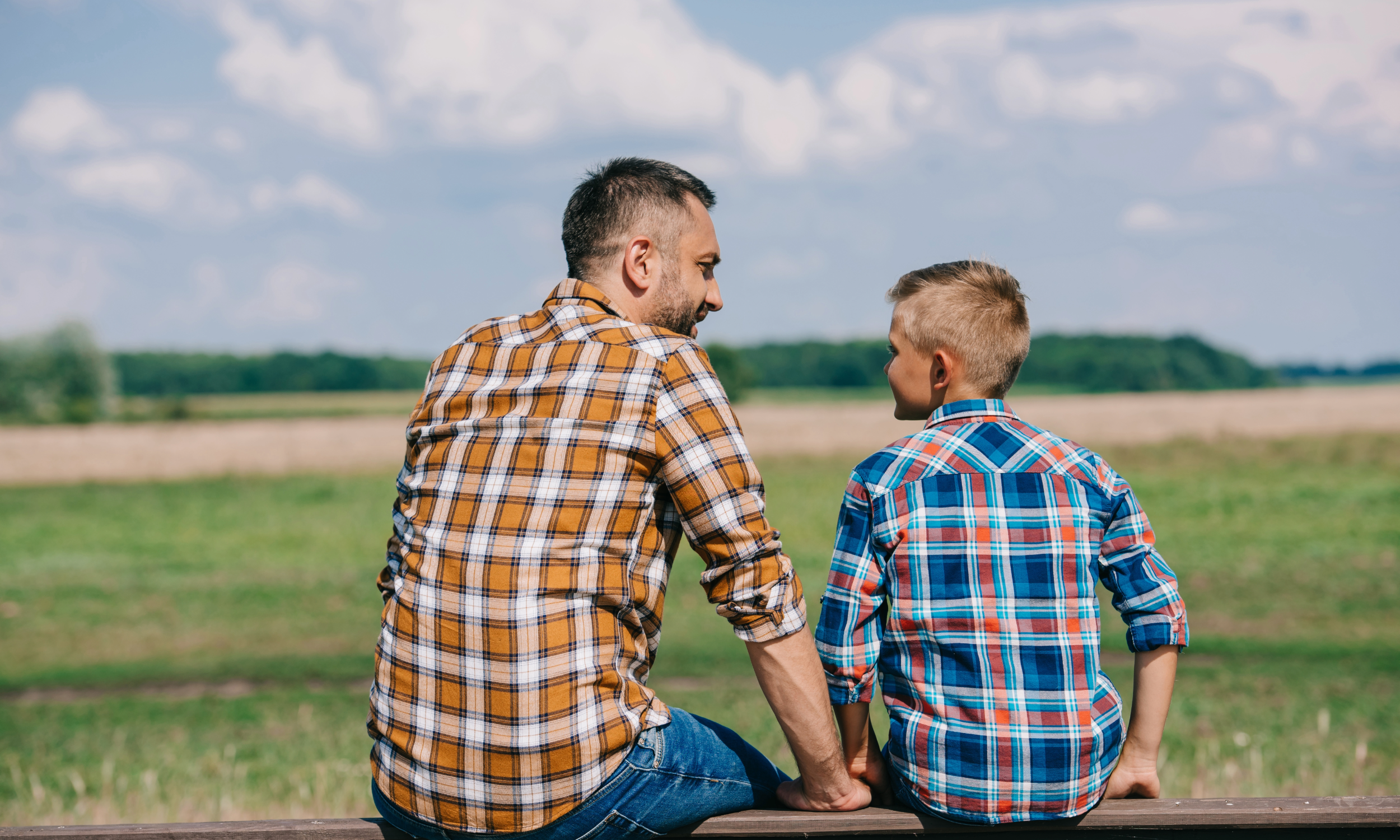 A man and a boy sitting on a bench outdoors, wearing plaid shirts, with a grassy field and blue sky with clouds in the background.