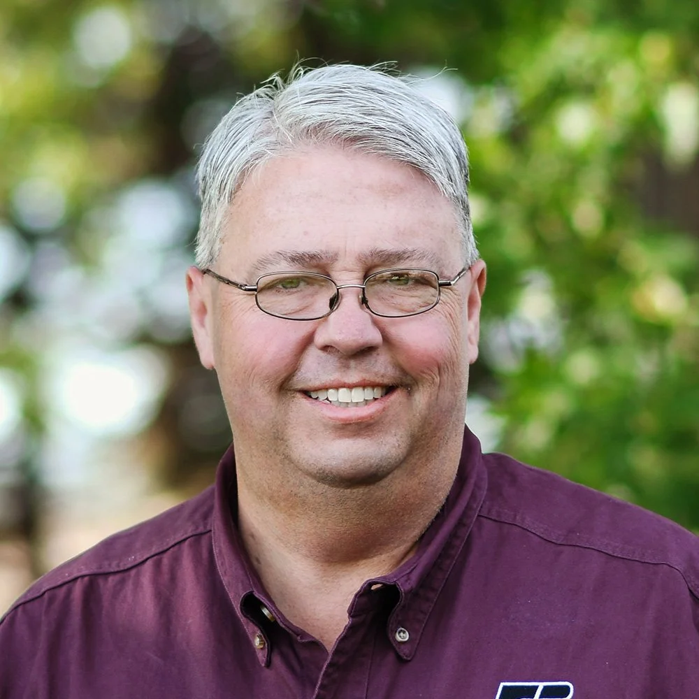 A middle-aged man with gray hair, wearing glasses and a maroon collared shirt, smiling outdoors with green blurred trees in the background.