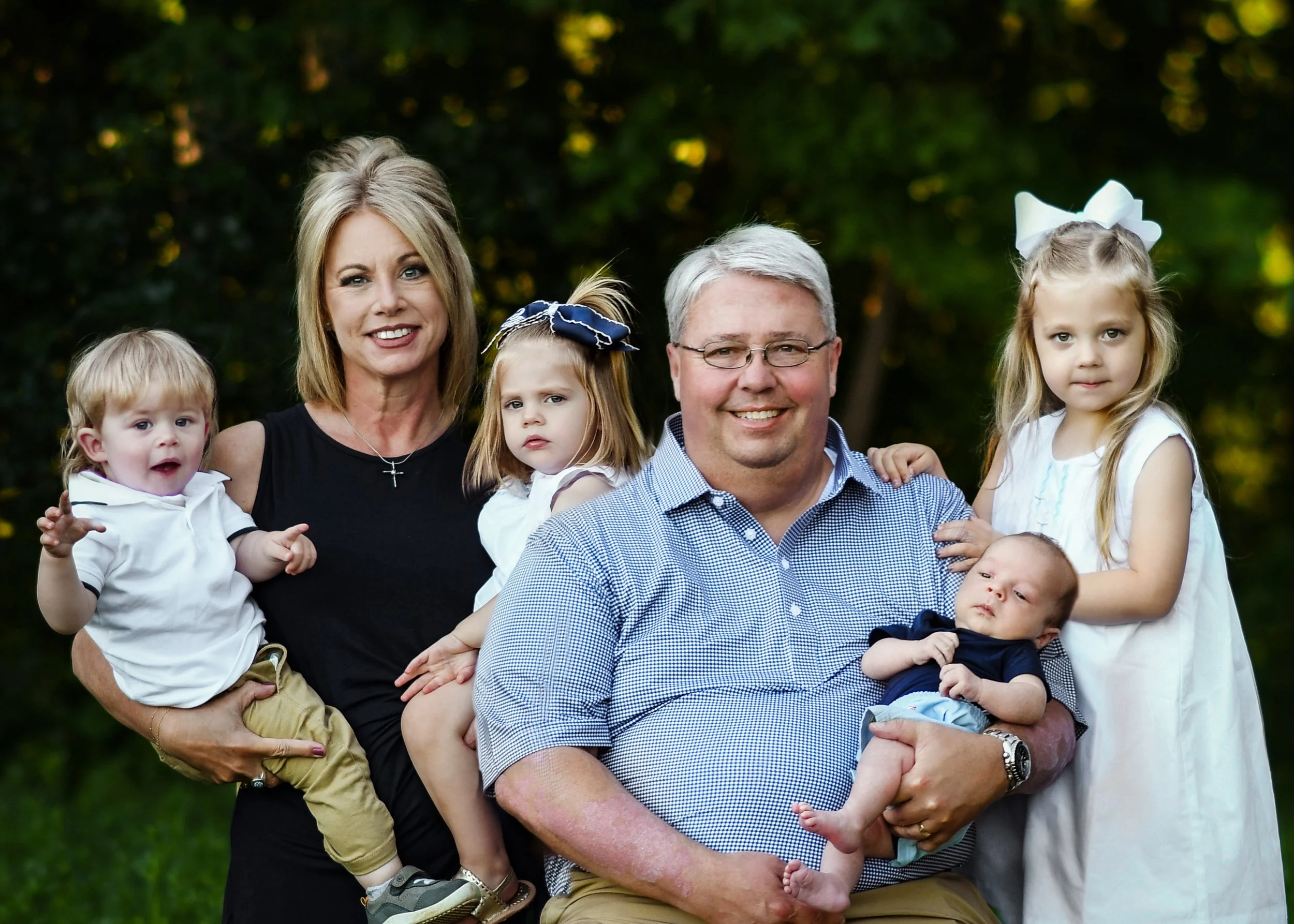 A large family group outdoors during daytime, consisting of two adults and five children, posing for a photo with green trees in the background.