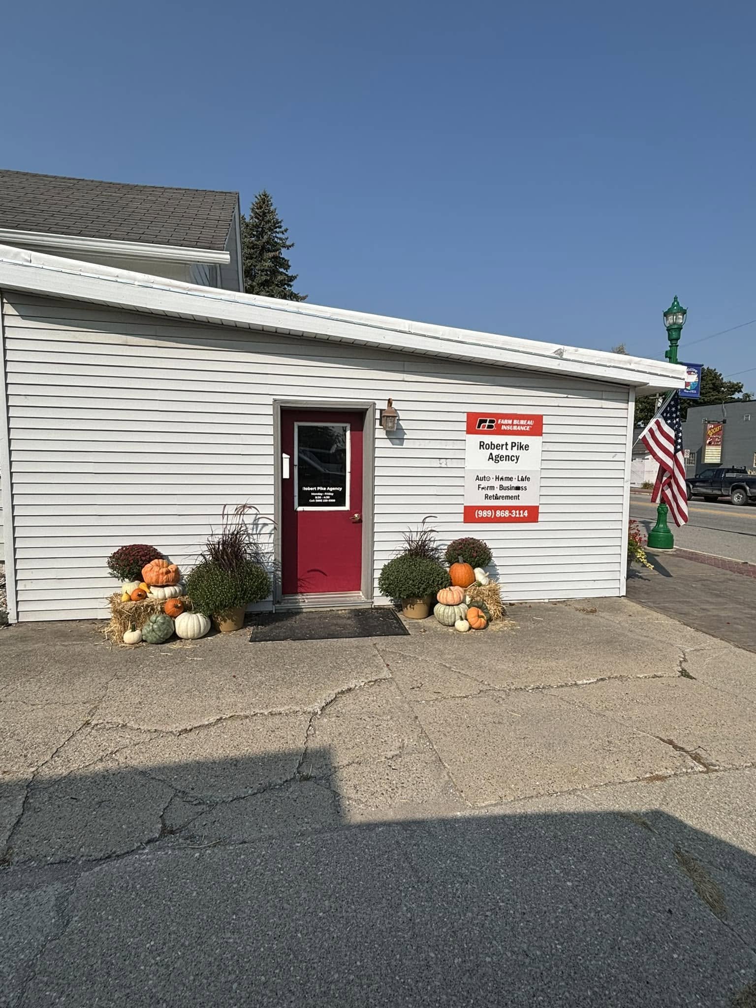 Small building with white siding, red door, and sign to the right. Clusters of pumpkins and fall decorations outside, American flag nearby. Clear blue sky.