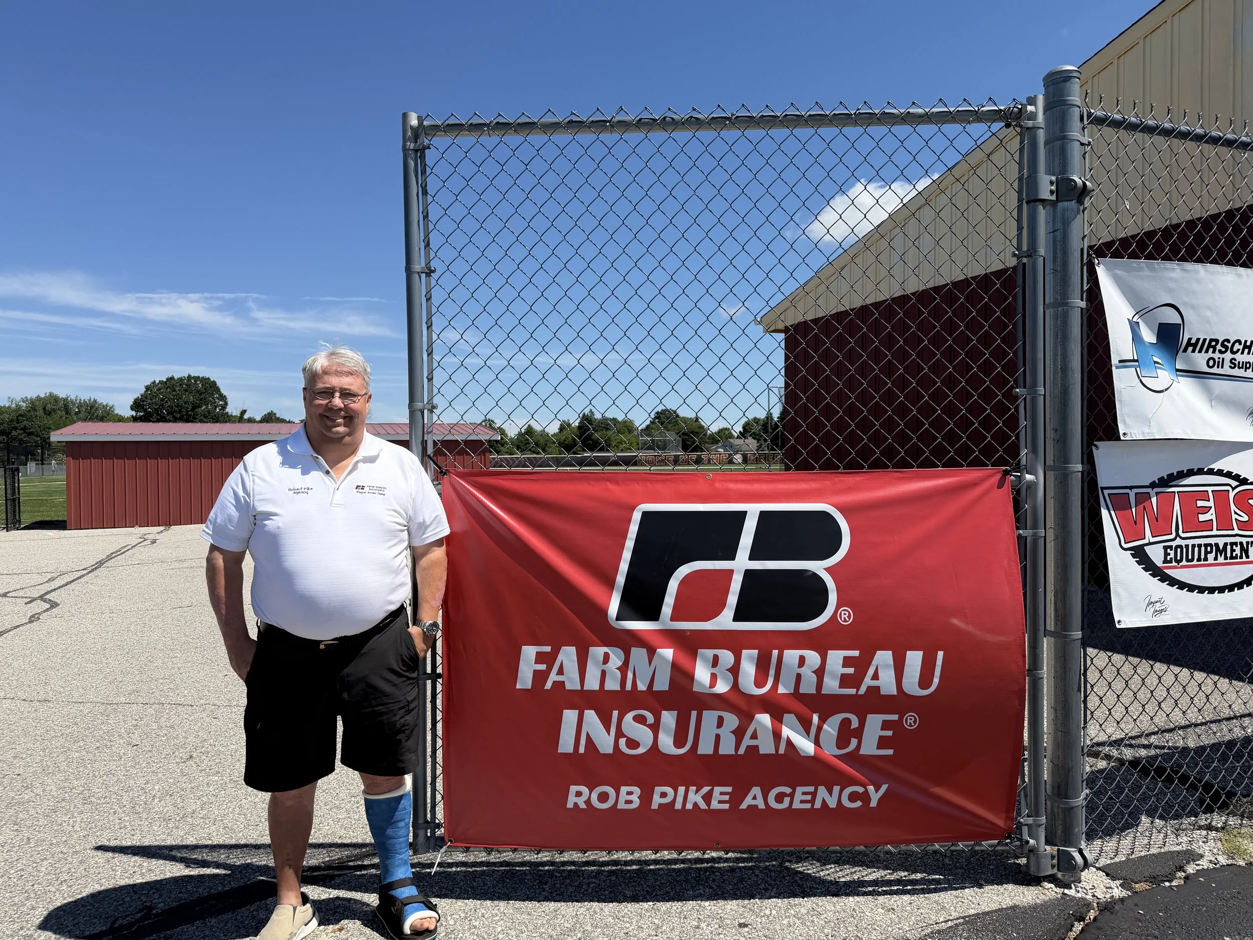 A man with white hair and glasses smiling, standing next to a person with a disabled leg with a cast, in front of a chain-link fence with a red banner that reads 'Farm Bureau Insurance' and a white banner with a logo.
