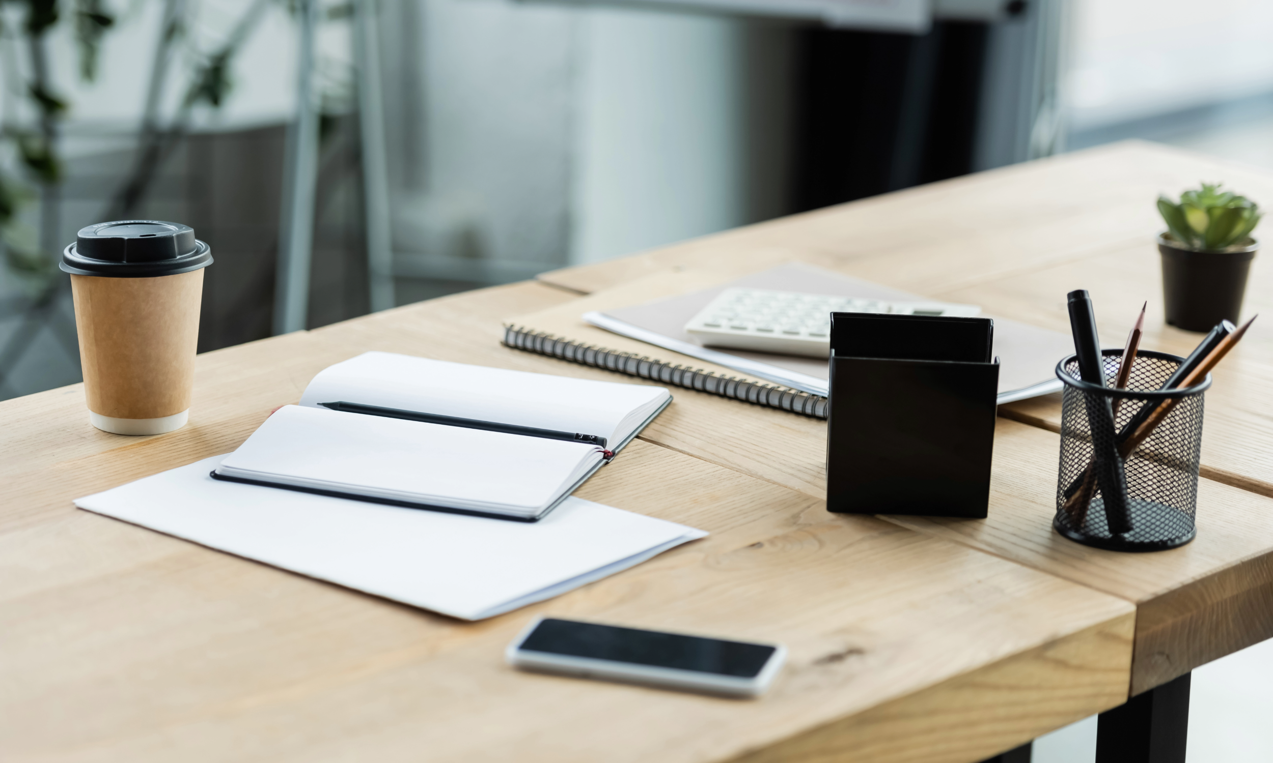 Office desk with a coffee cup, open notebook, pen, calculator, closed notebook, smartphone, pen holder with pencils, and a small potted plant.