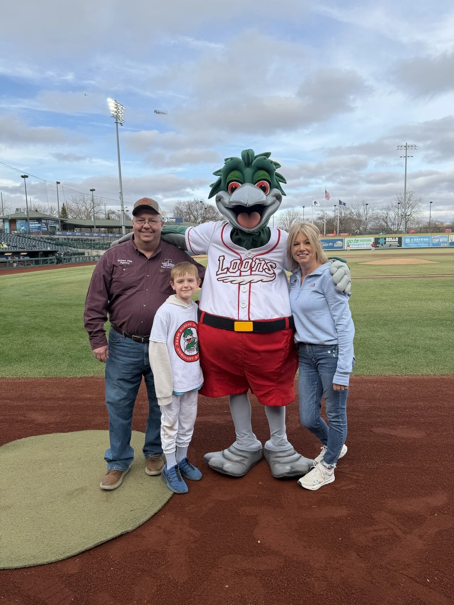 Three people and a baseball mascot posing on a baseball field, with a mostly cloudy sky and empty stadium seats in the background.