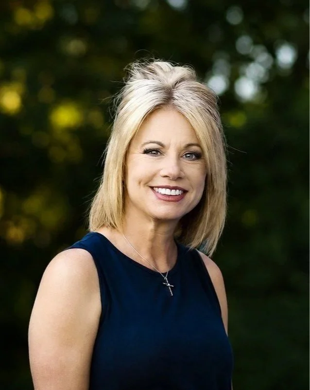 A smiling woman with blonde hair in a sleeveless navy blue top outdoors, with blurred green trees in the background.