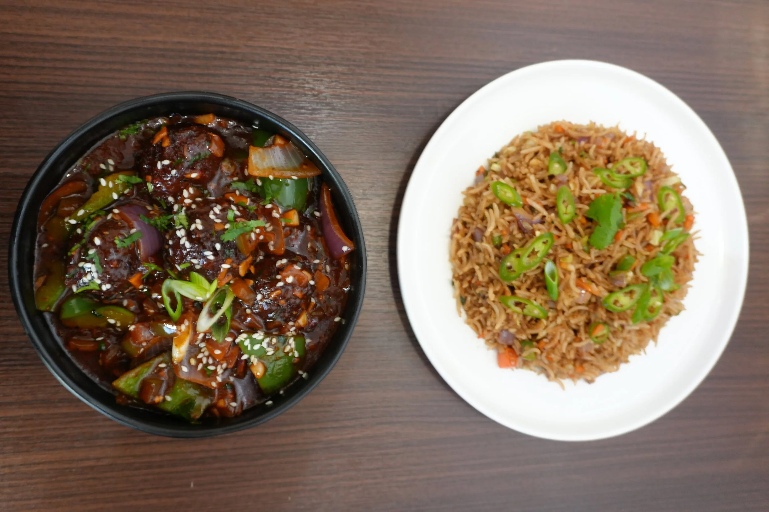 A black bowl of beef stir-fry with vegetables and sesame seeds and a white plate of fried rice garnished with sliced green chili peppers and cilantro on a wooden table.