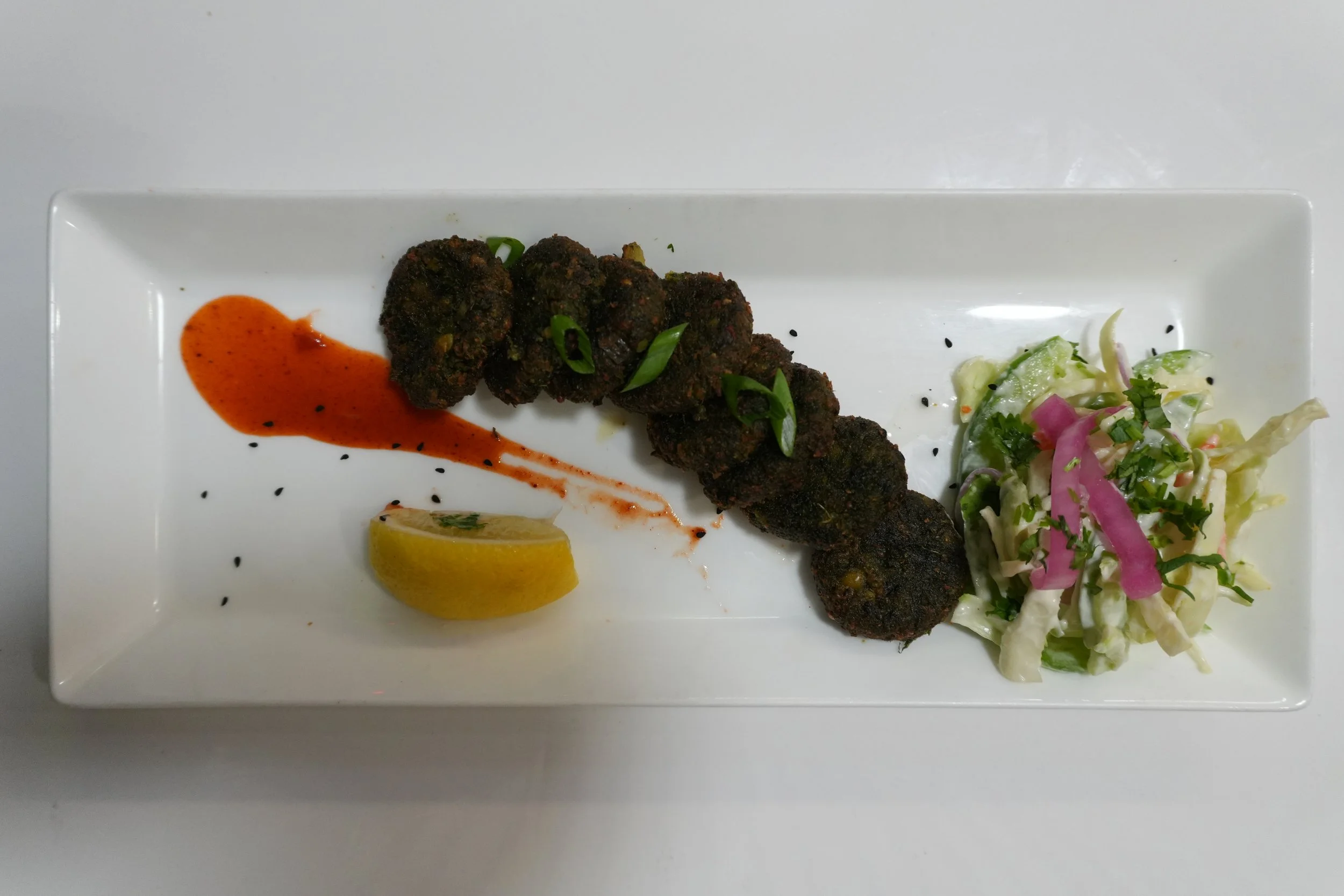 Plate with four fried croquettes garnished with chopped green onions, a lemon wedge, a drizzle of red sauce, and a side salad with cabbage, pickled onions, and herbs.