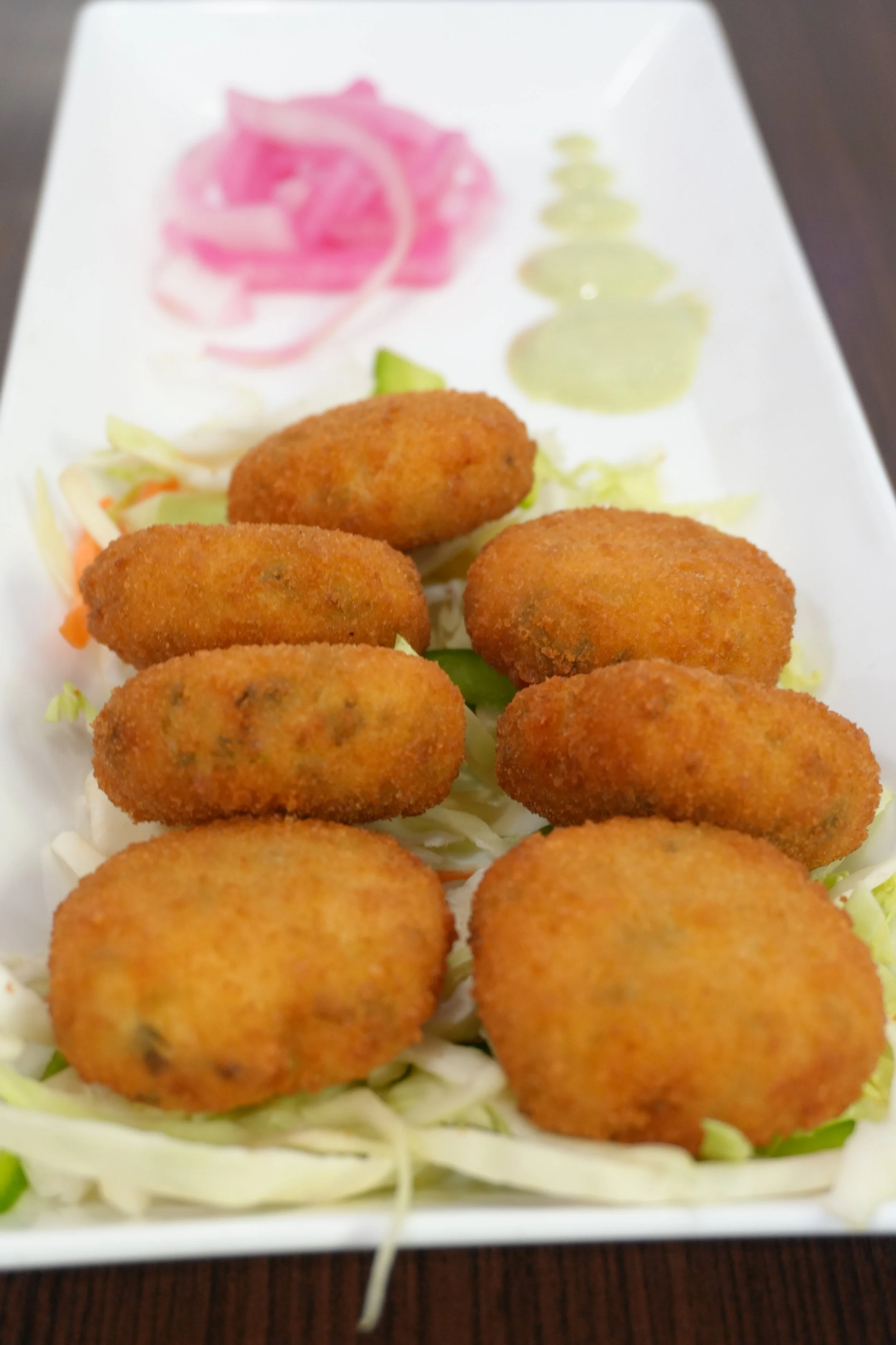Fried croquettes served on a bed of shredded cabbage, with pickled ginger and thinly sliced cucumber on a white rectangular plate.