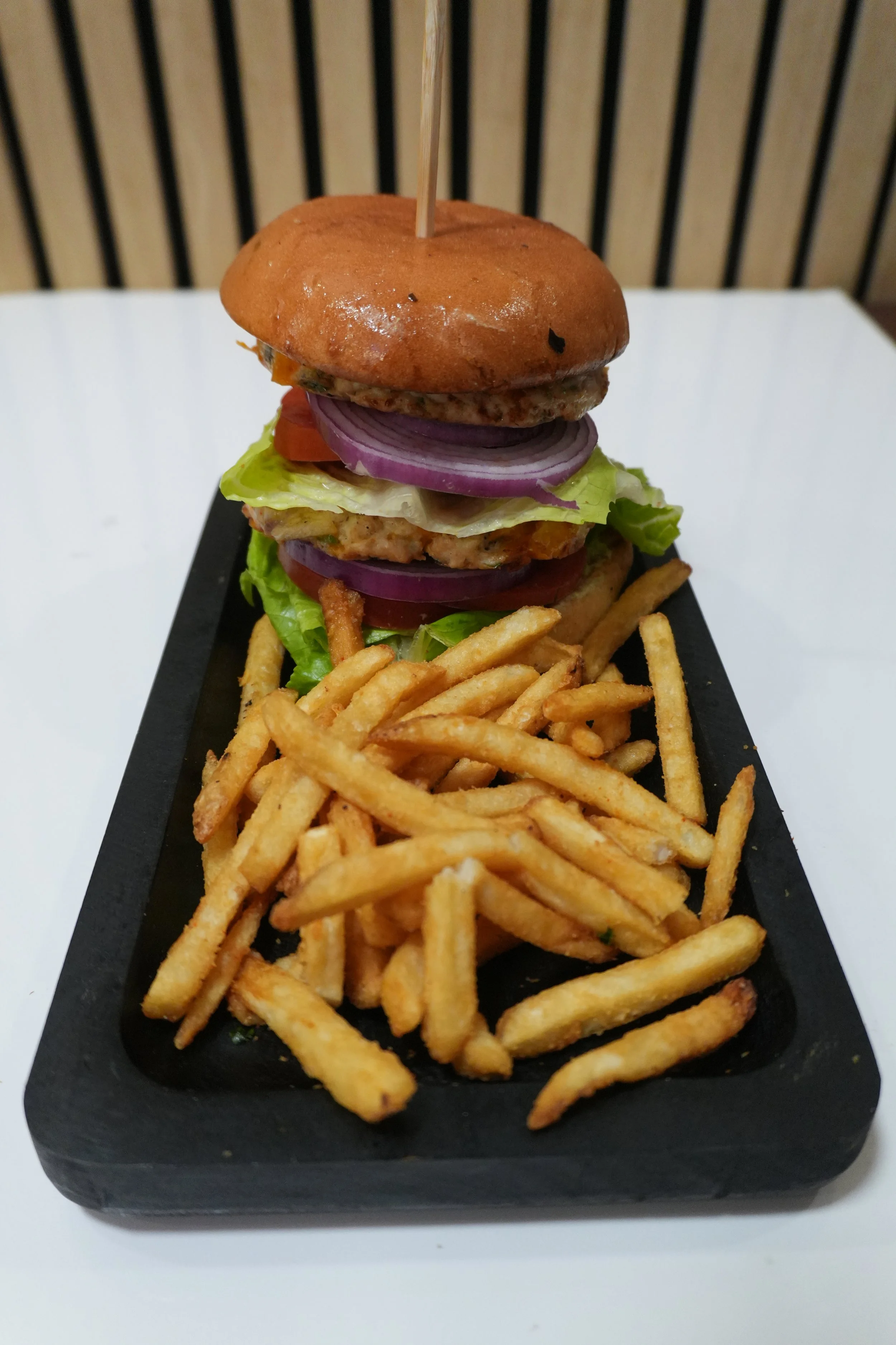 A burger with lettuce, tomato, onions, a burger patty, cheese, and a bun, served with fries on a black serving tray.