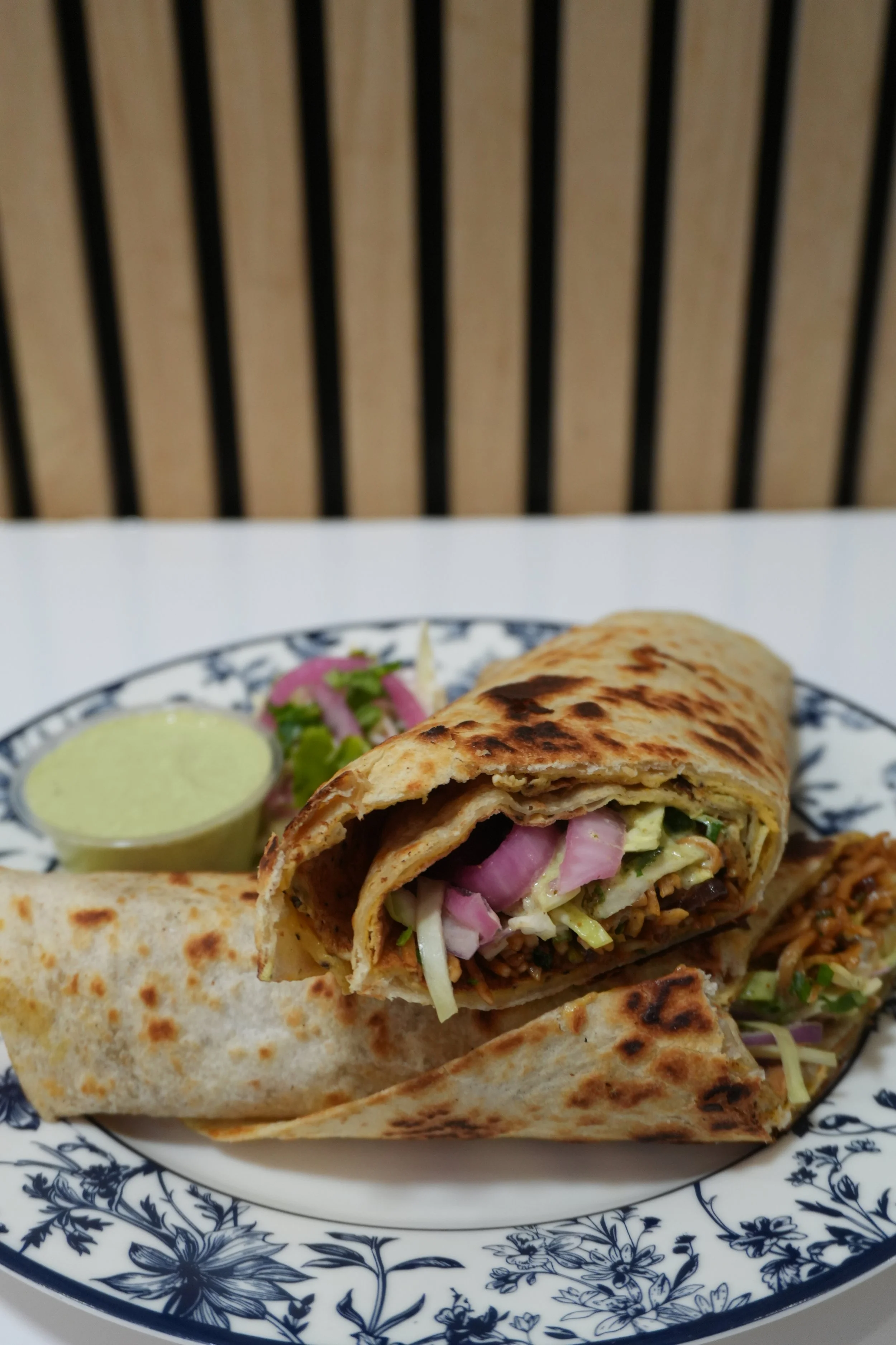 Close-up of a burrito cut in half on a decorative plate with a side of green sauce and some pickled vegetables, with a wooden slat background.