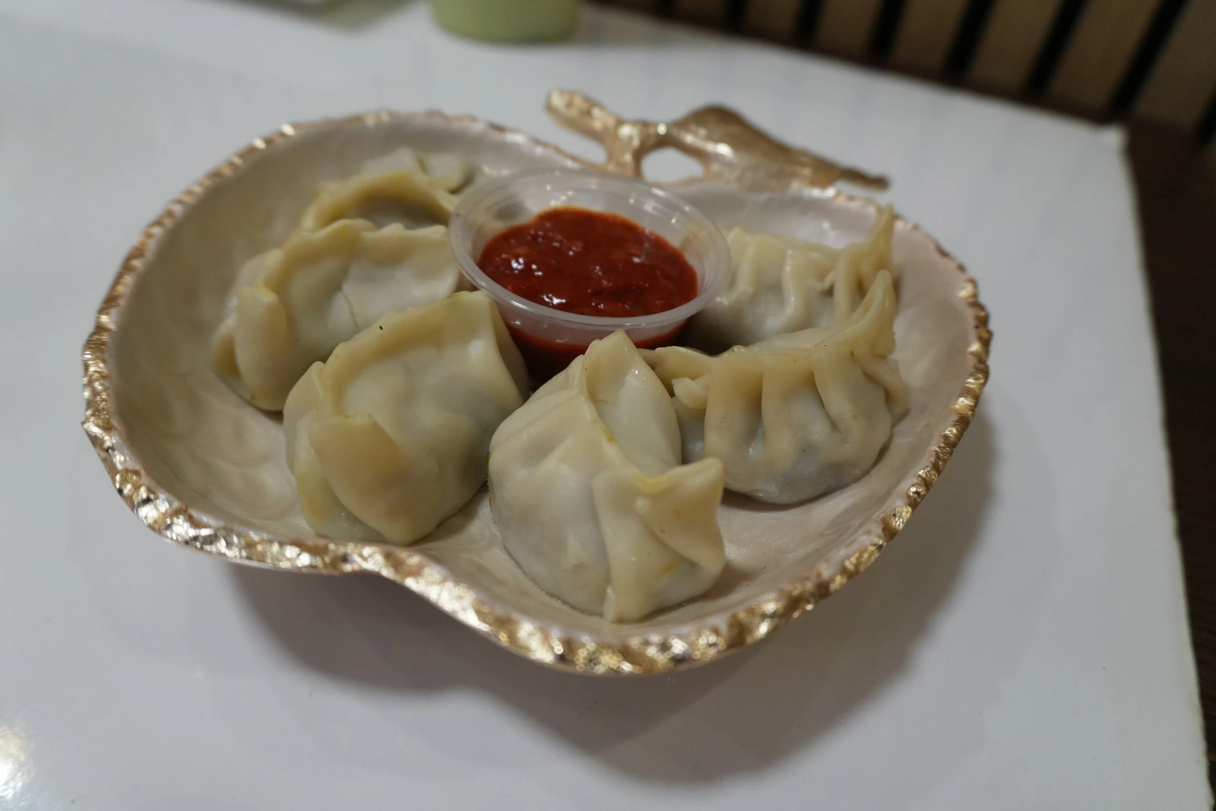 A small round dish with a gold rim filled with steamed dumplings, topped with a small cup of red dipping sauce.