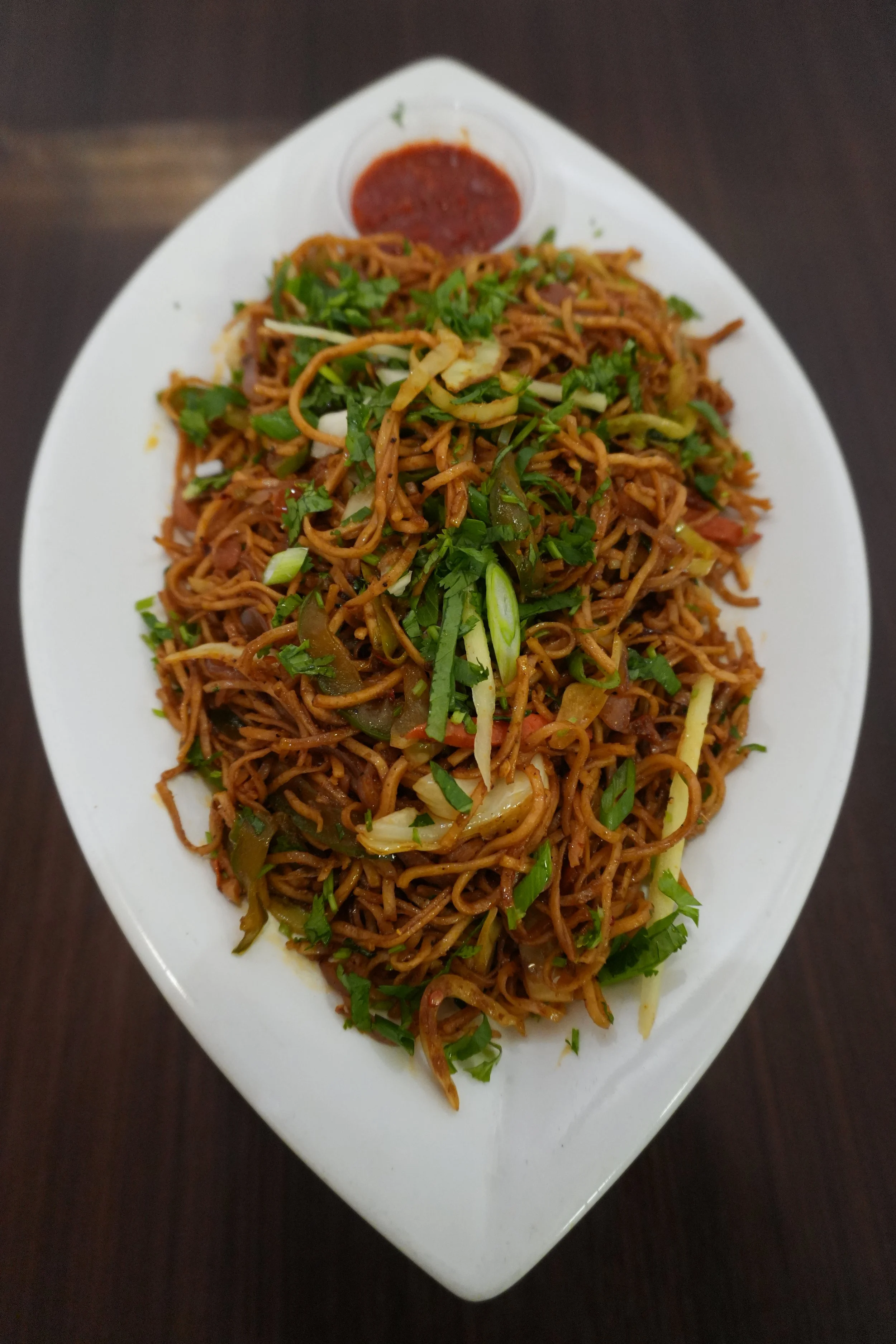 Plate of stir-fried noodles with vegetables garnished with chopped cilantro, served with spicy sauce on the side.