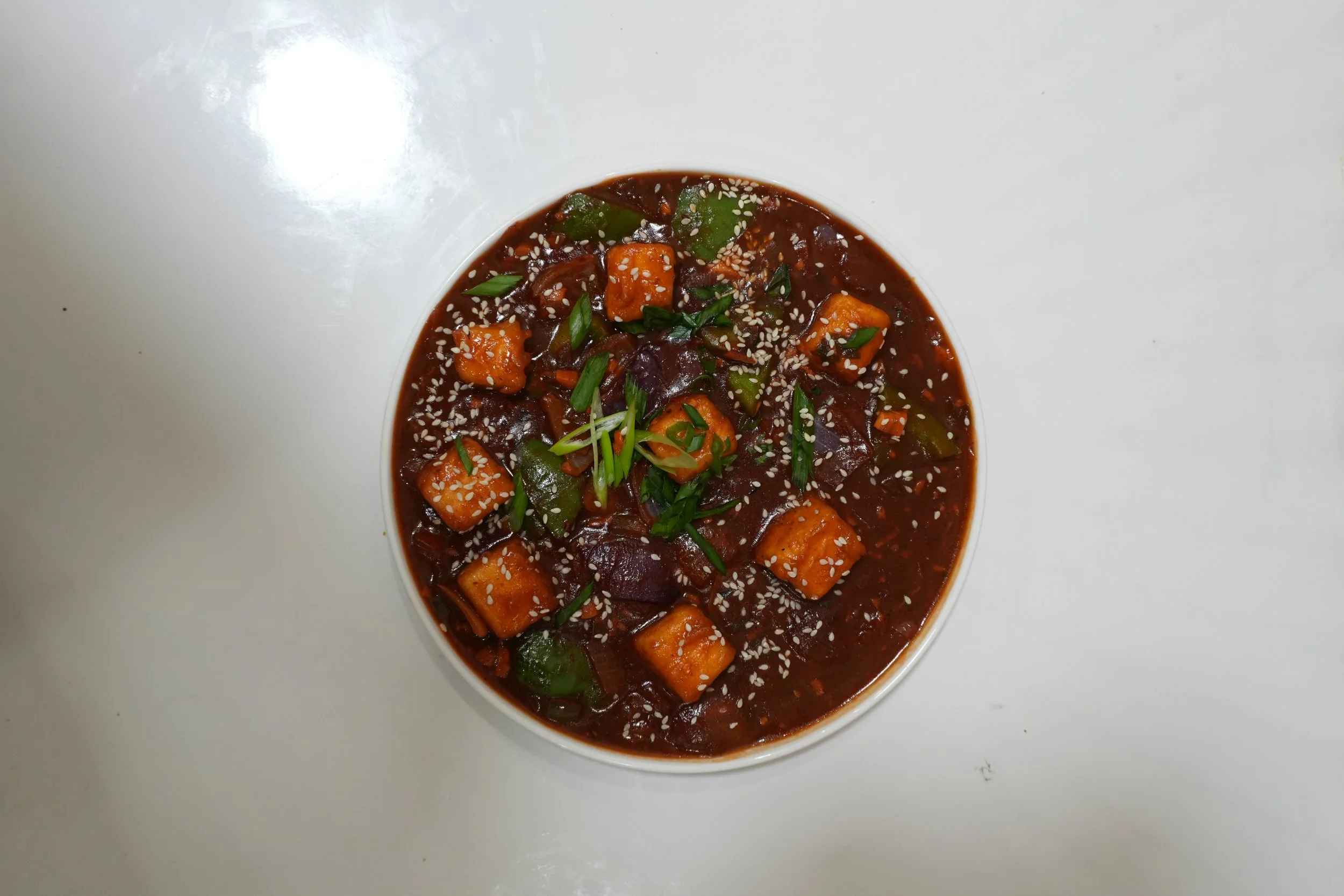 A bowl of Korean spicy stew with diced vegetables, possibly sweet potato and zucchini, garnished with sesame seeds and green onions, on a white surface.