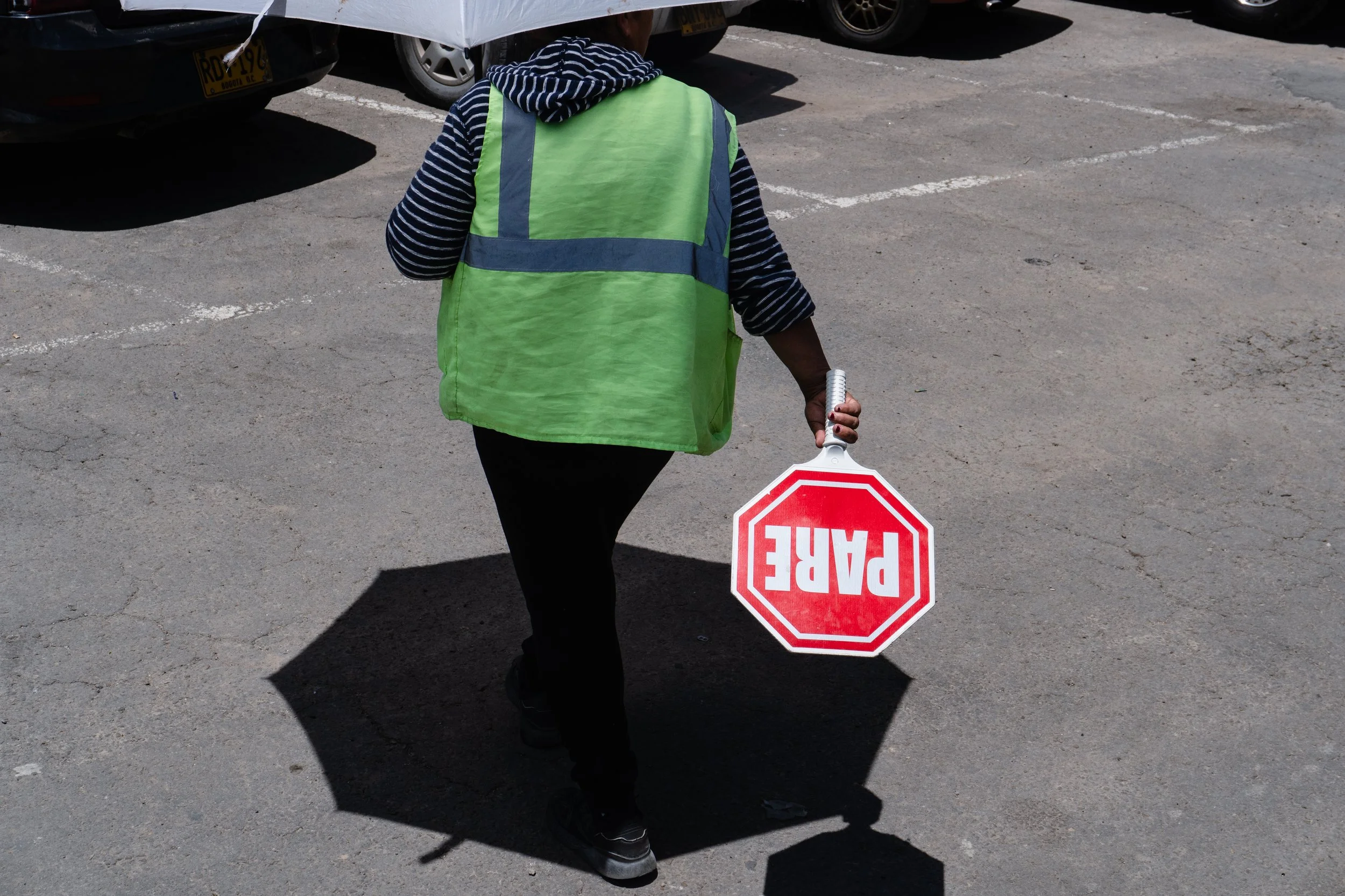 Person in a green safety vest holding a stop sign while walking on a parking lot under sunny weather.