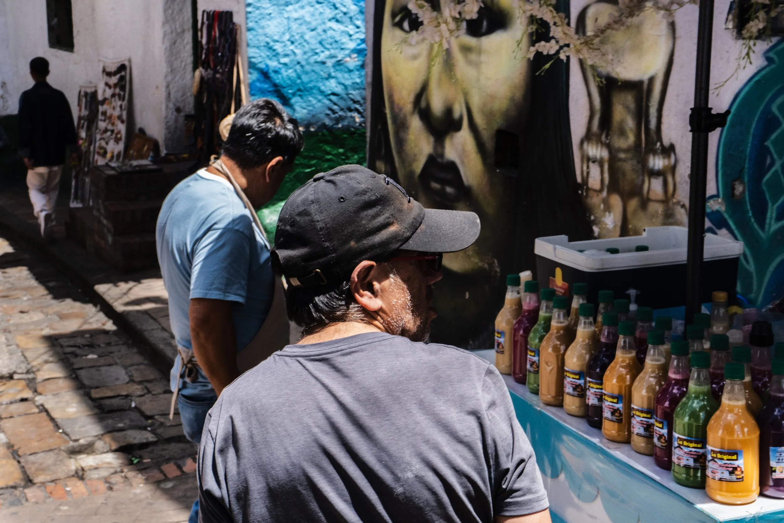 Two men browsing bottles of colorful beverages on a street vendor cart, with vibrant street art mural in the background.