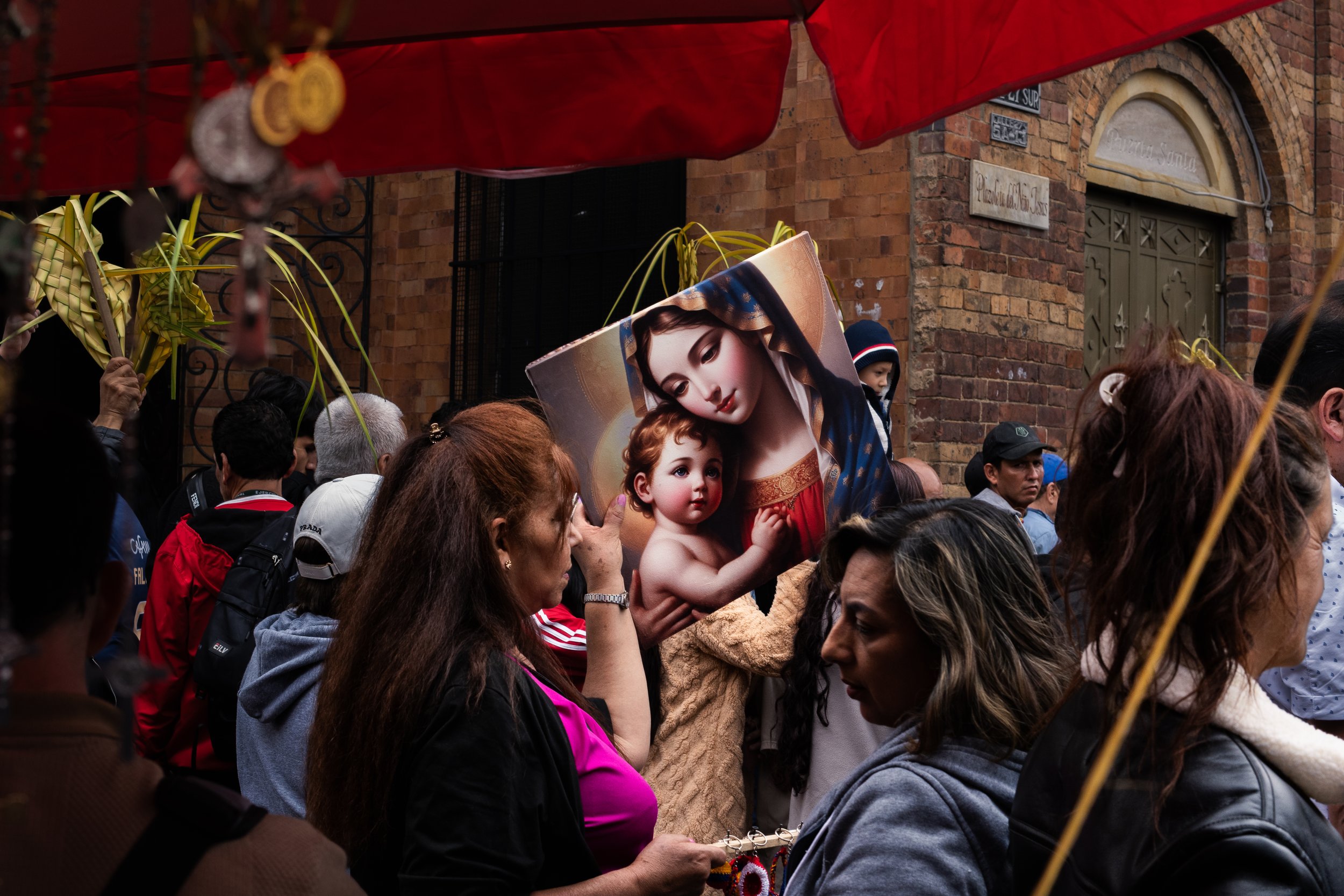 A group of people gathered outdoors, some holding yellow palm leaves and a picture of Mary and baby Jesus, against a brick building with a green door.