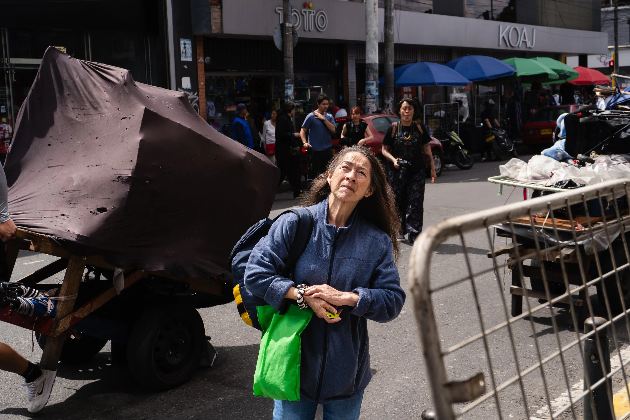 An elderly woman in a blue jacket with a green bag on her arm stands on a busy city street, looking upward. Behind her, there are people, umbrellas, and street vendors, with stores in the background.