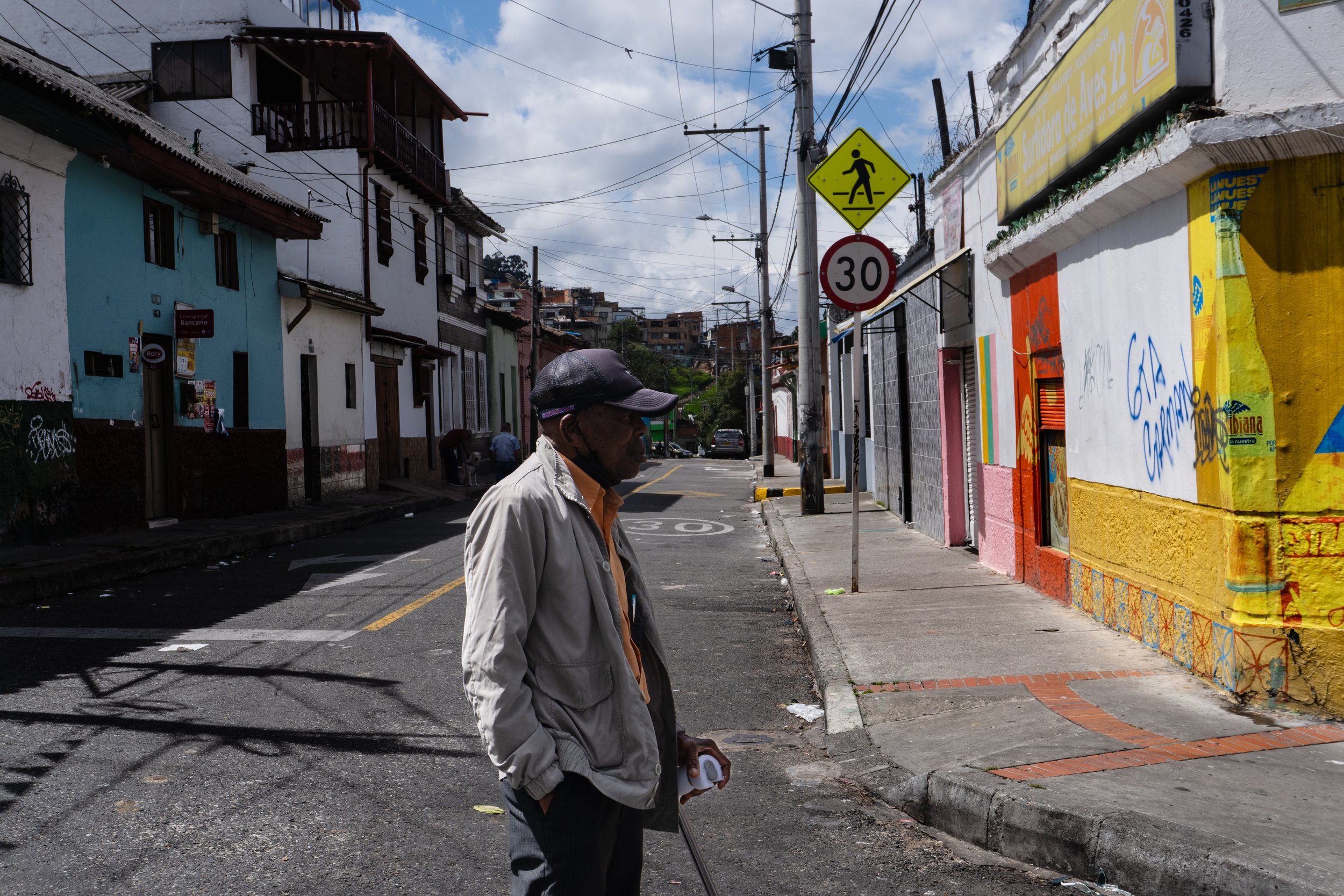A man in a beige jacket and black hat standing on a sidewalk in a colorful urban neighborhood, with graffiti on the building wall, street signs, and houses in the background.
