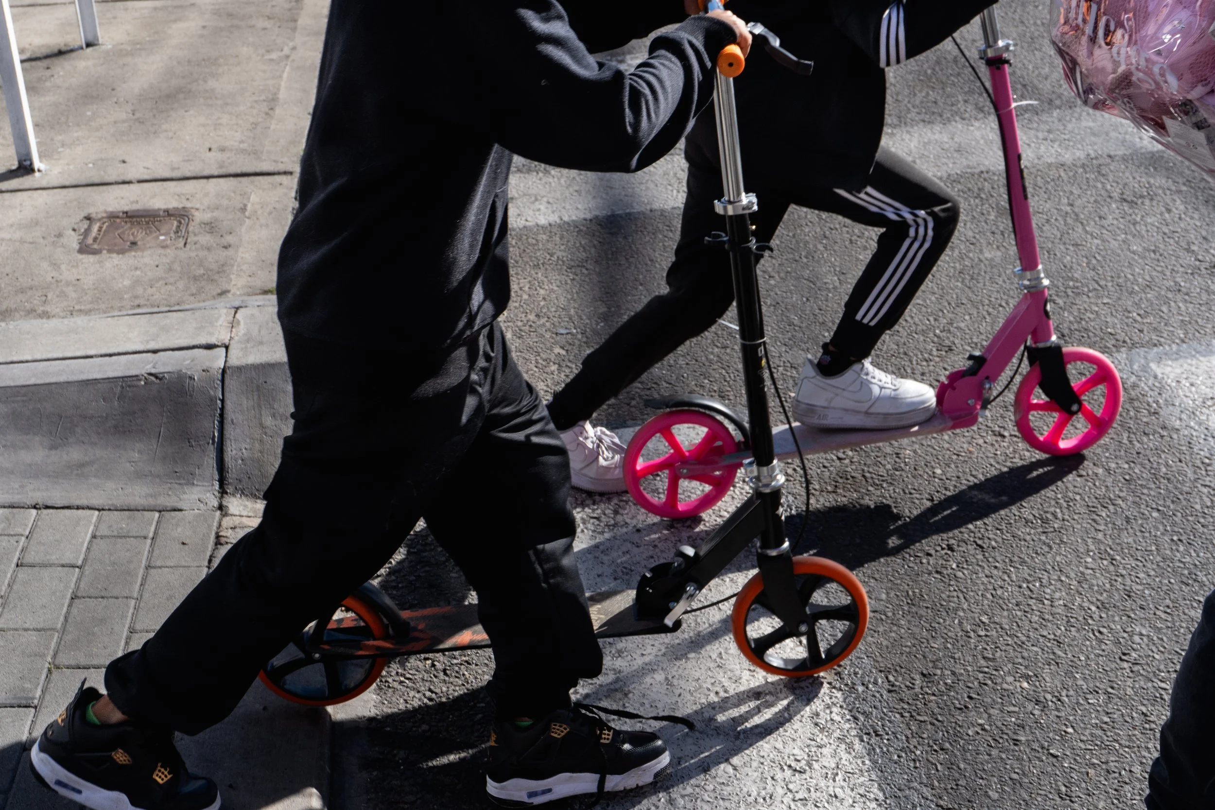 Two kids riding scooters on a city street, one with a black and orange scooter and the other on a pink and white scooter, both dressed in black athletic clothing.
