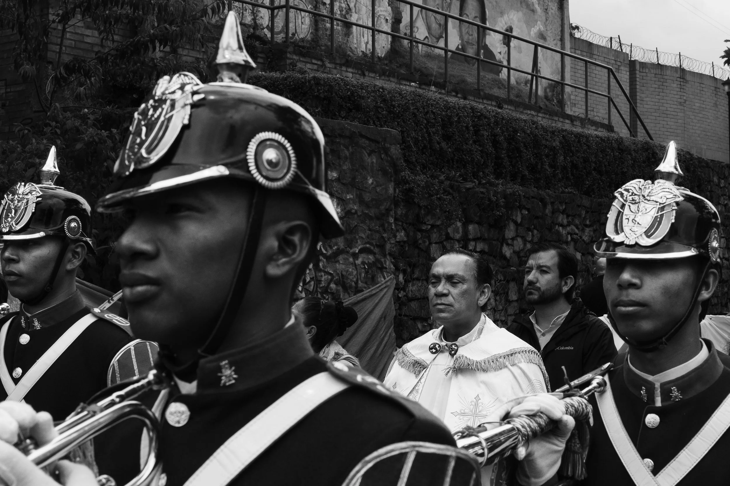 A black and white photo of uniformed soldiers in a marching band, with two men standing behind them in traditional clothing, possibly during a cultural ceremony or parade.
