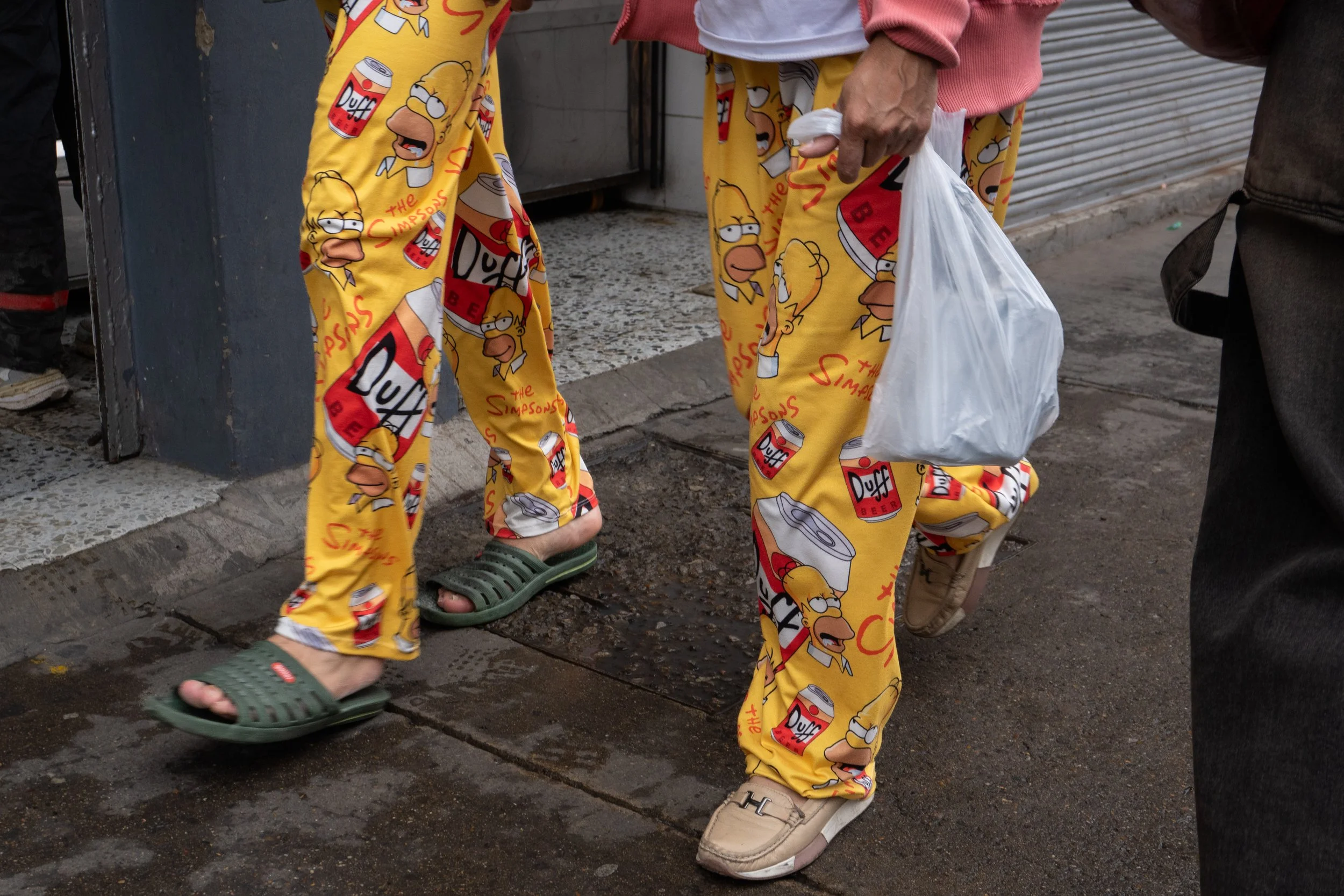 Two people wearing yellow pajama pants with Simpsons characters and Duff beer cans, standing on a wet sidewalk. One person is wearing green slide sandals; the other is in beige sneakers and holding a plastic bag.