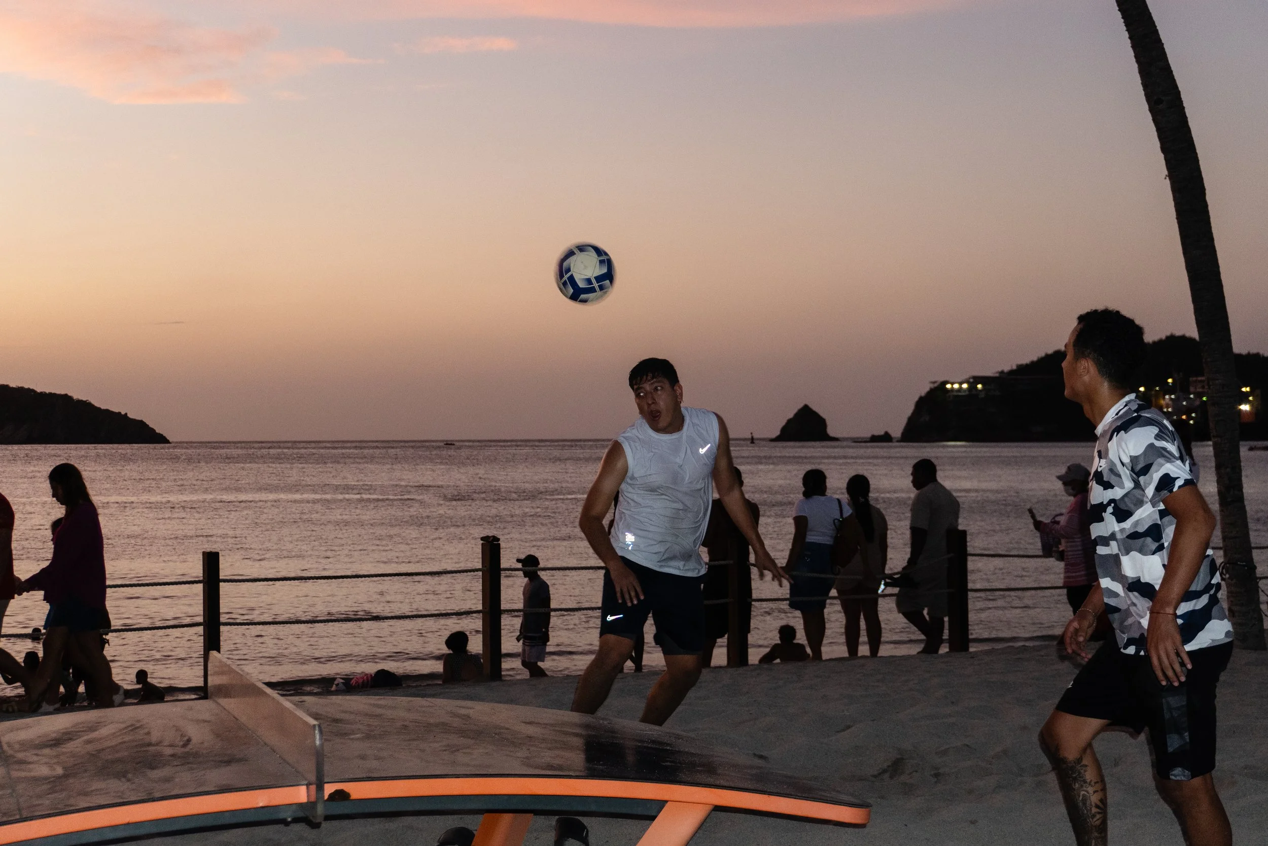 People playing volleyball on the beach at sunset, with the ocean and distant cliffs in the background.