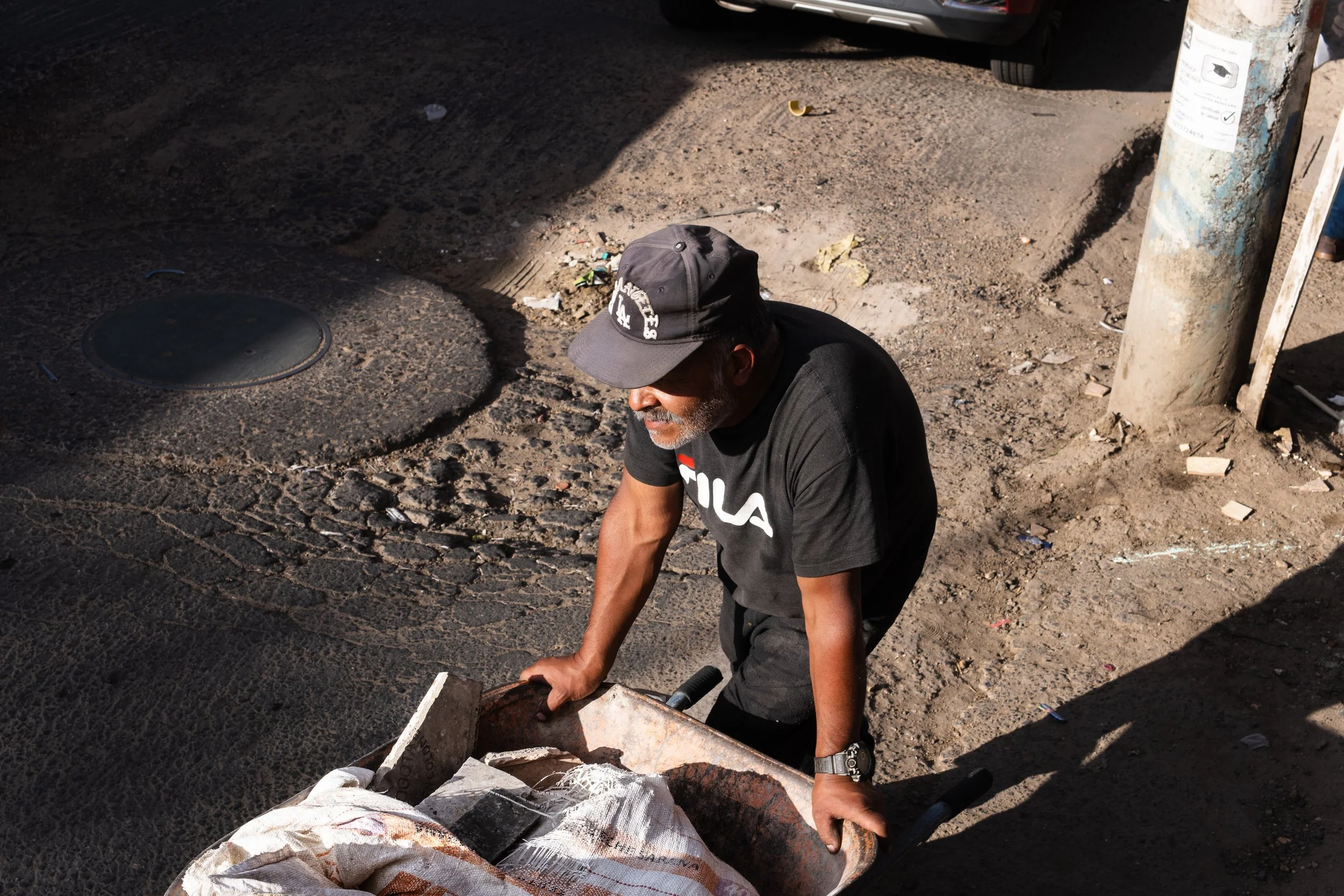 A man wearing a black FILA t-shirt and a black cap pushing a rusty wheelbarrow on a dirt and asphalt street. The street has some debris and a manhole cover. Sunlight and shadows are visible.