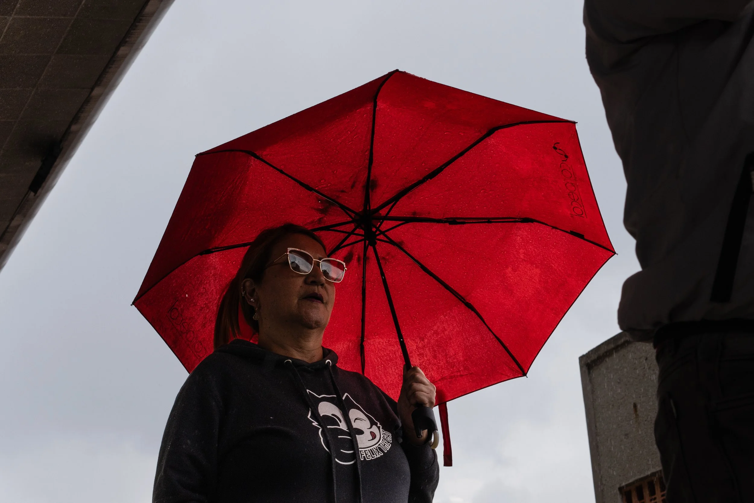 A woman holding a red umbrella outdoors, with rain droplets on the umbrella, wearing sunglasses and a hoodie, with an overcast sky in the background.