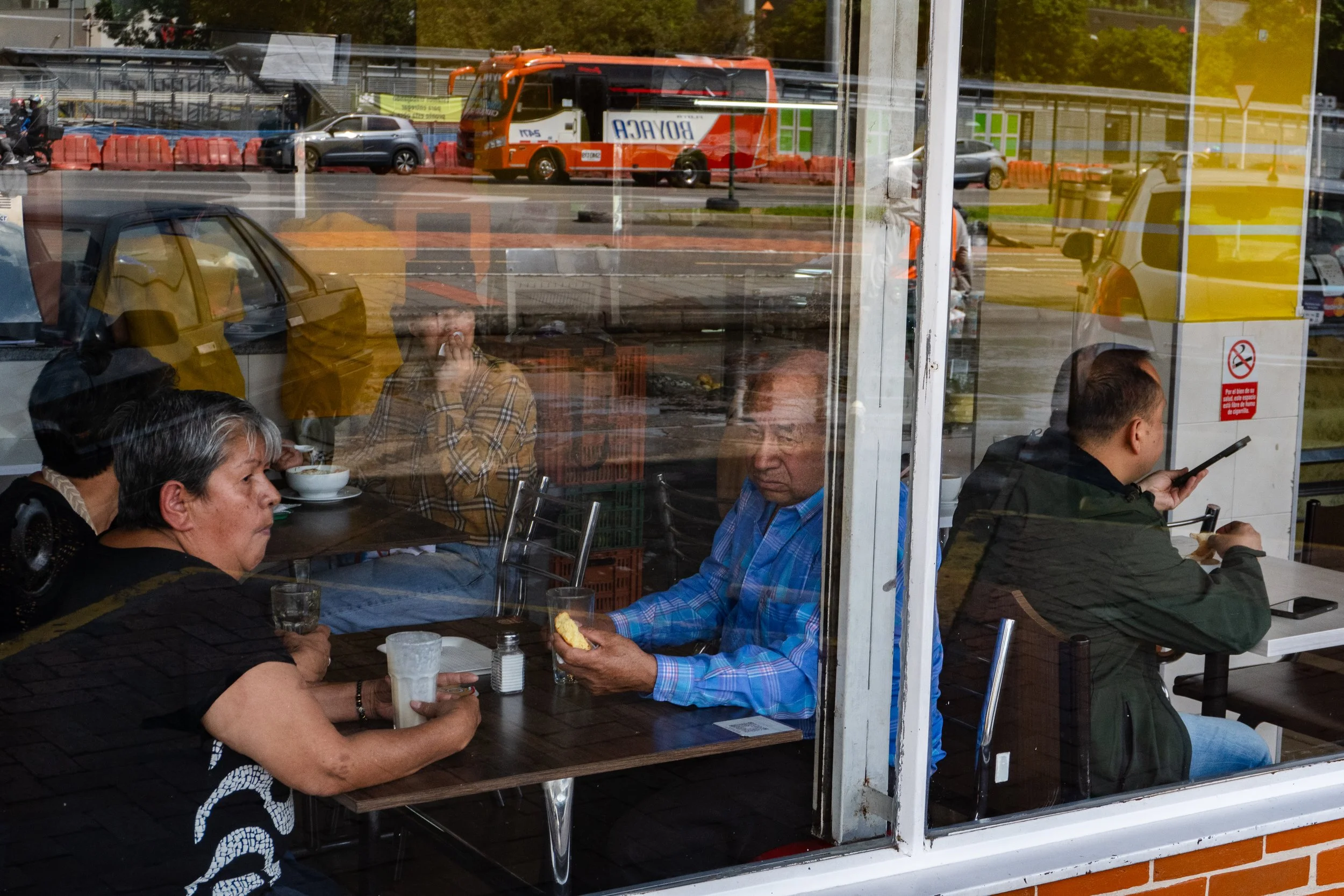 People dining inside a restaurant, viewed through a glass window, with street traffic and a bus outside.