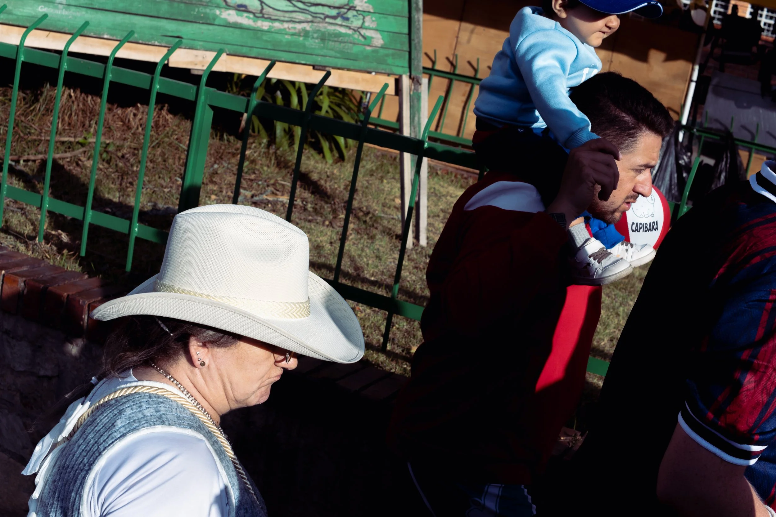 A woman wearing a white cowboy hat and glasses, a man carrying a child on his shoulders at an outdoor event, a green fence, and part of a sports setting with a rugby ball labeled "Capibara."