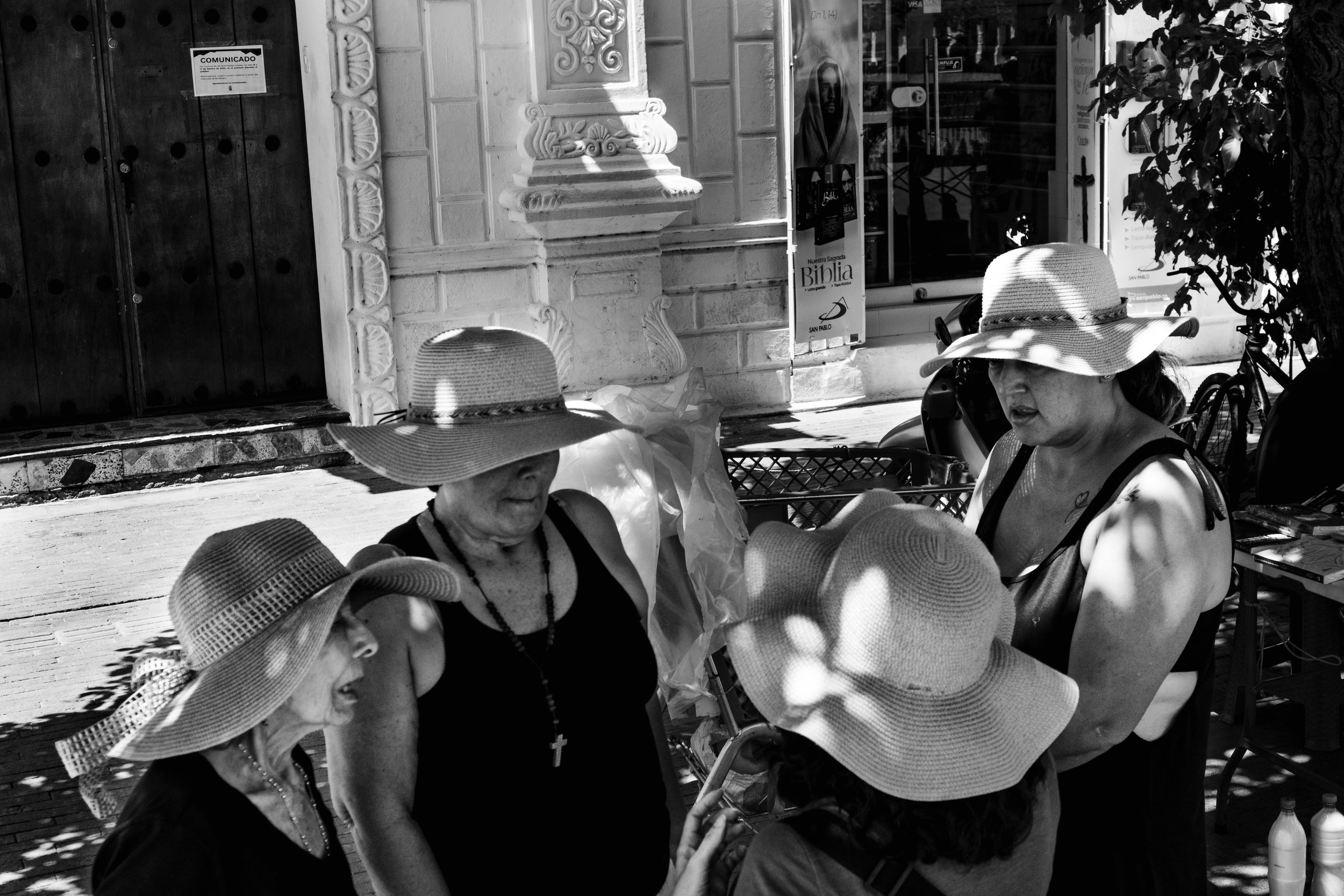 A group of five women, all wearing large brimmed sun hats, are standing together outdoors, talking and smiling in shaded sunlight.