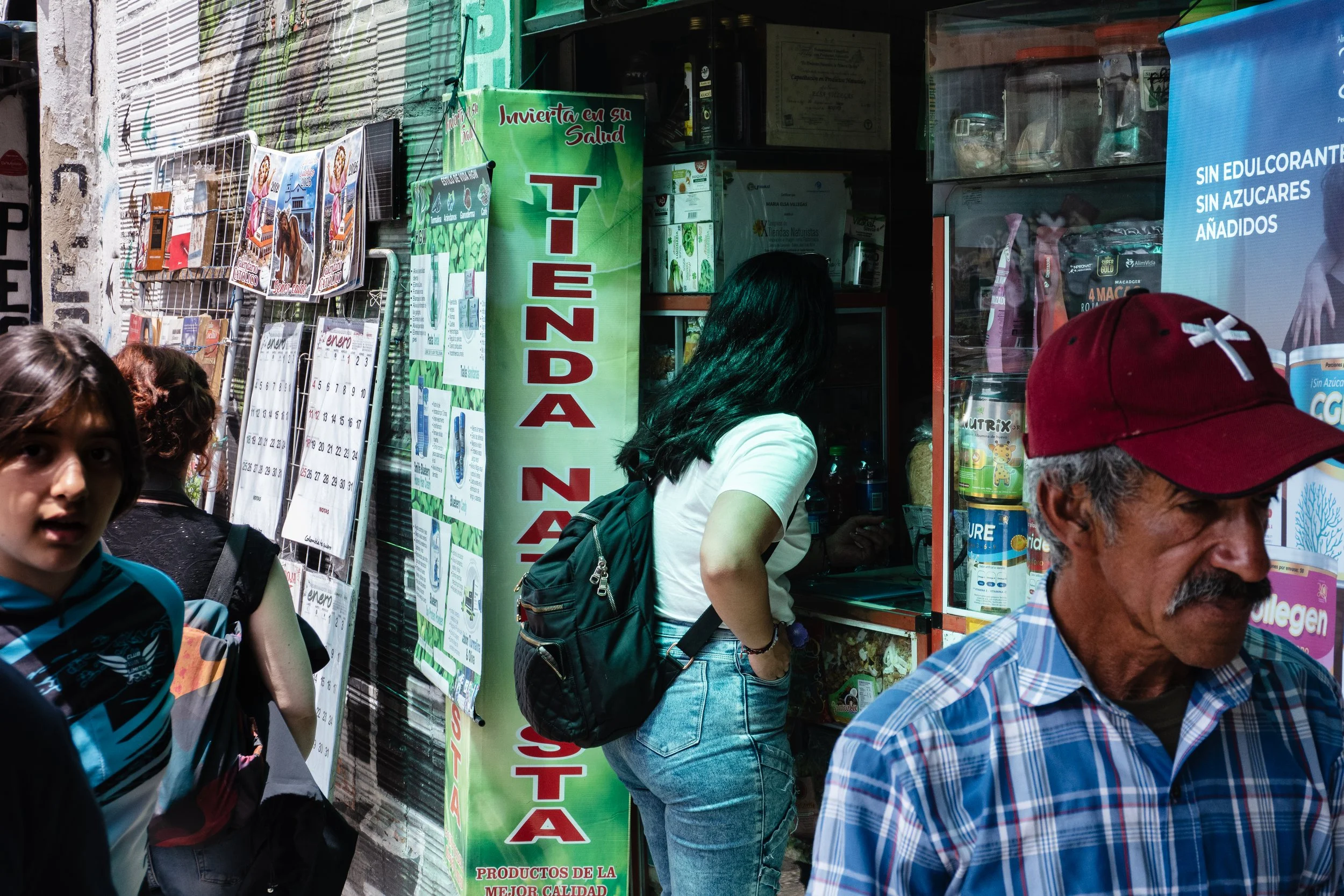 People shopping outside a small store with a green sign that says 'Tienda' and various posters and advertisements displayed on the wall.