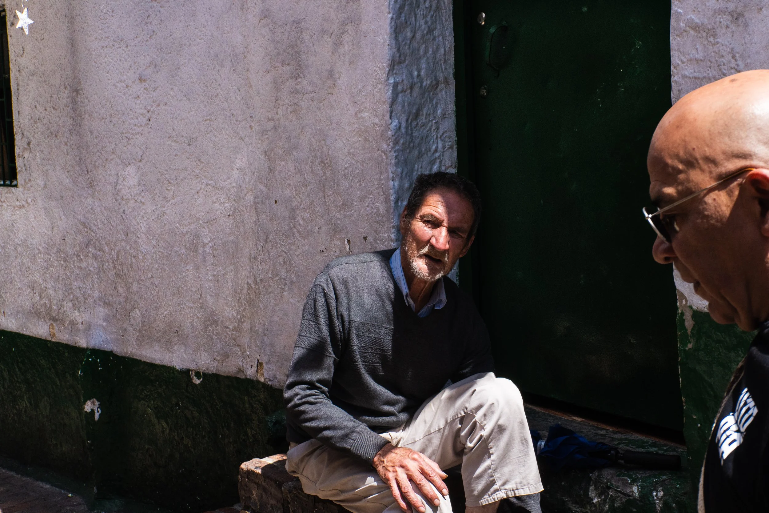 Two men are outside against a weathered wall. One man is sitting on a small bench, looking at the camera, while the other man is standing nearby, turned to the side and partially out of the frame.