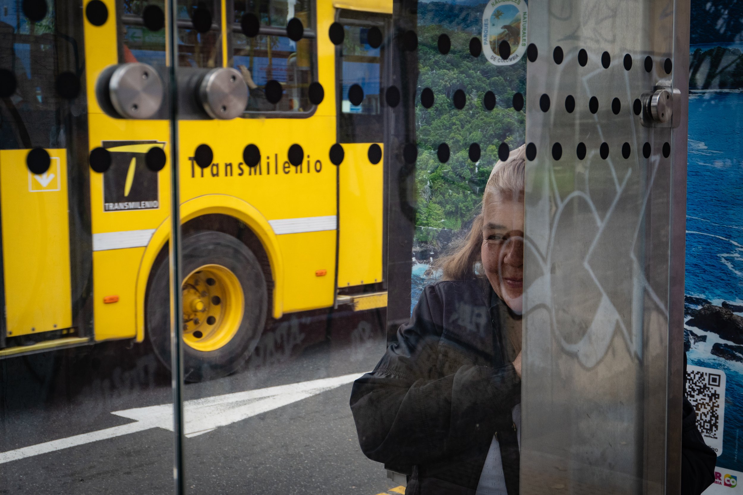 A reflection of a woman smiling and wearing glasses, seen through a glass door or window with black dots, with a yellow bus labeled 'Transmilenio' visible outside and a scenic landscape of the ocean and greenery in the background.