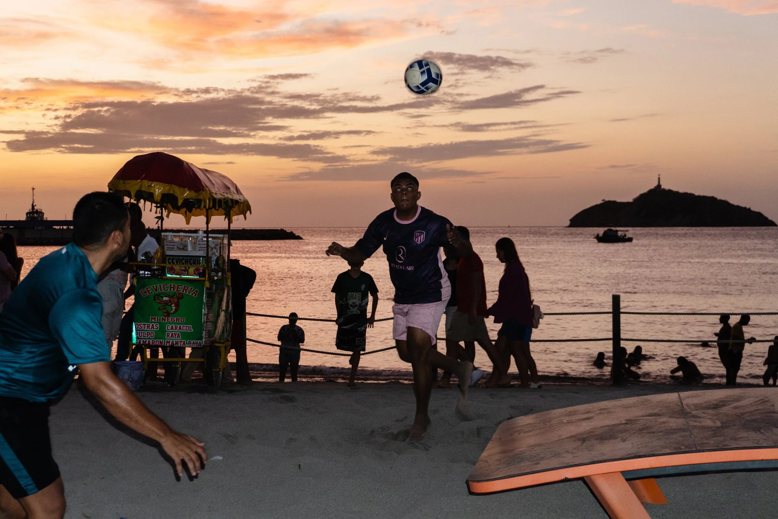 People playing beach volleyball at sunset on a sandy beach, with an island and boats visible in the background.
